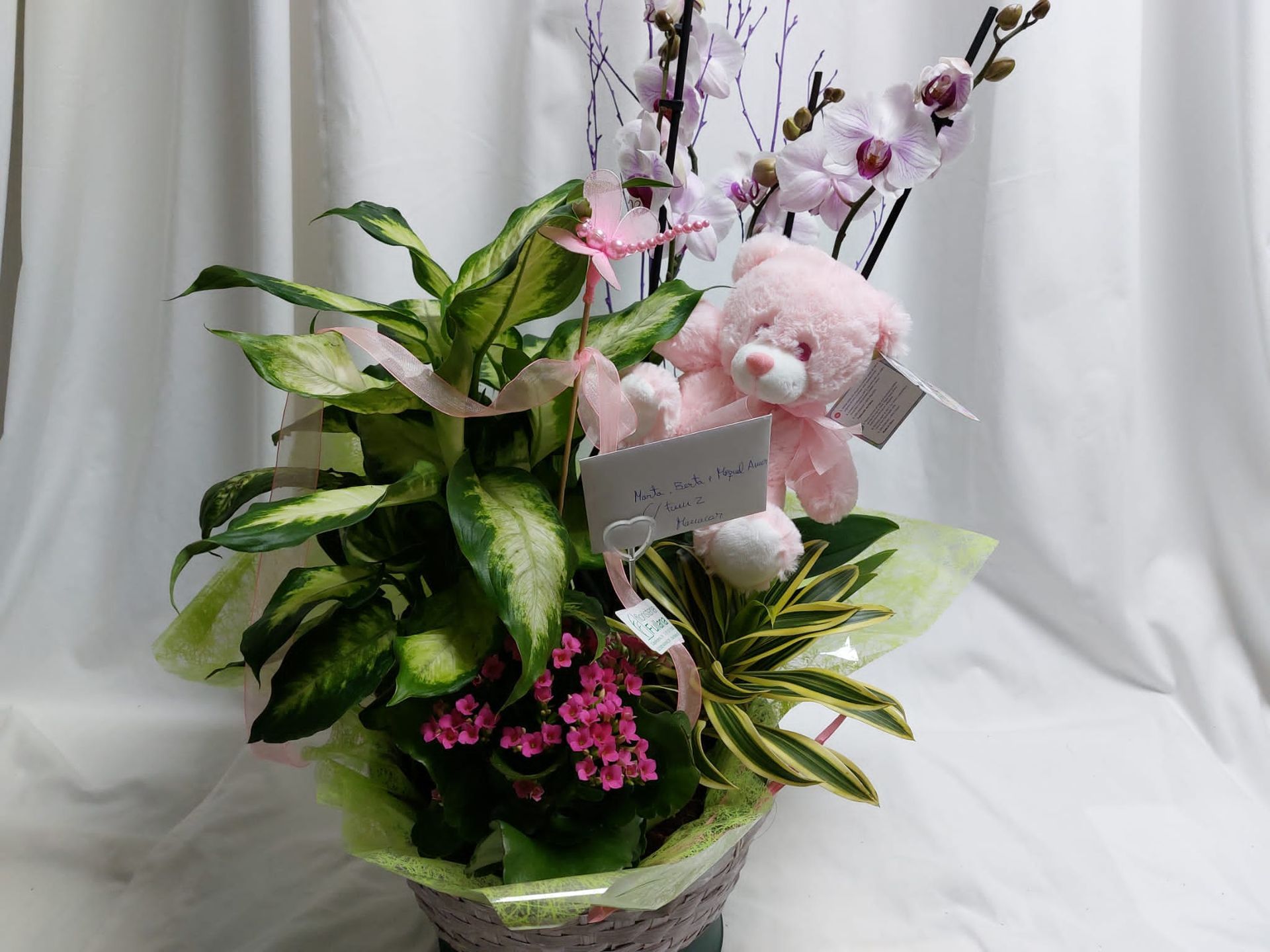Un arreglo floral en una canasta con orquídeas rosas, follaje verde, un osito de peluche rosa y pequeñas flores rosas.