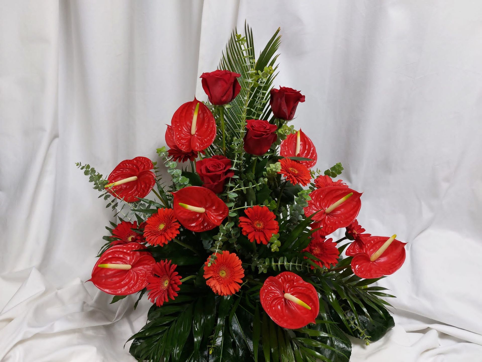 Un arreglo floral con anturios rojos, rosas y margaritas gerberas, sobre un fondo blanco.