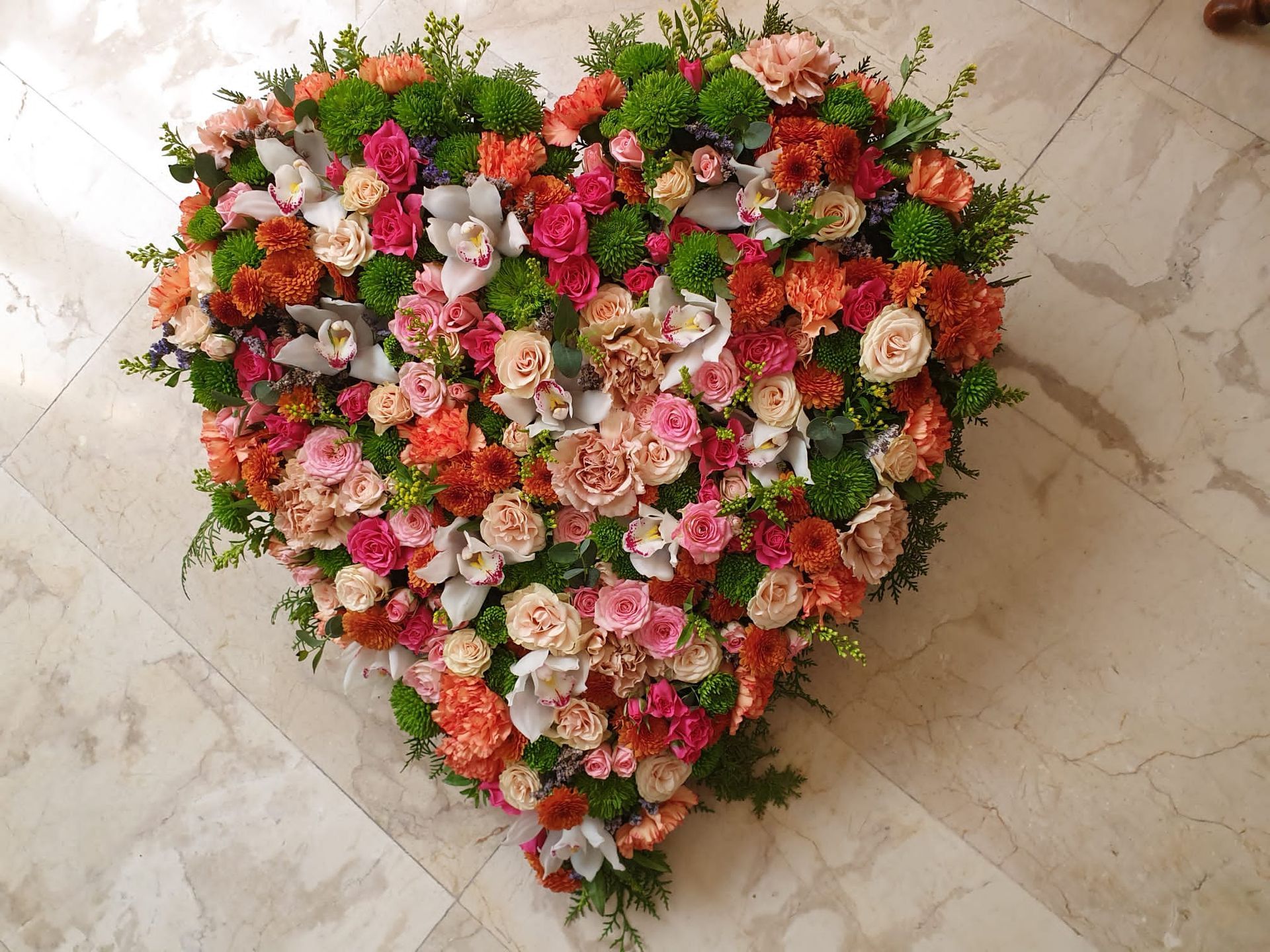 Arreglo floral en forma de corazón en tonos rosa, naranja y verde, apoyado sobre un suelo de mármol.