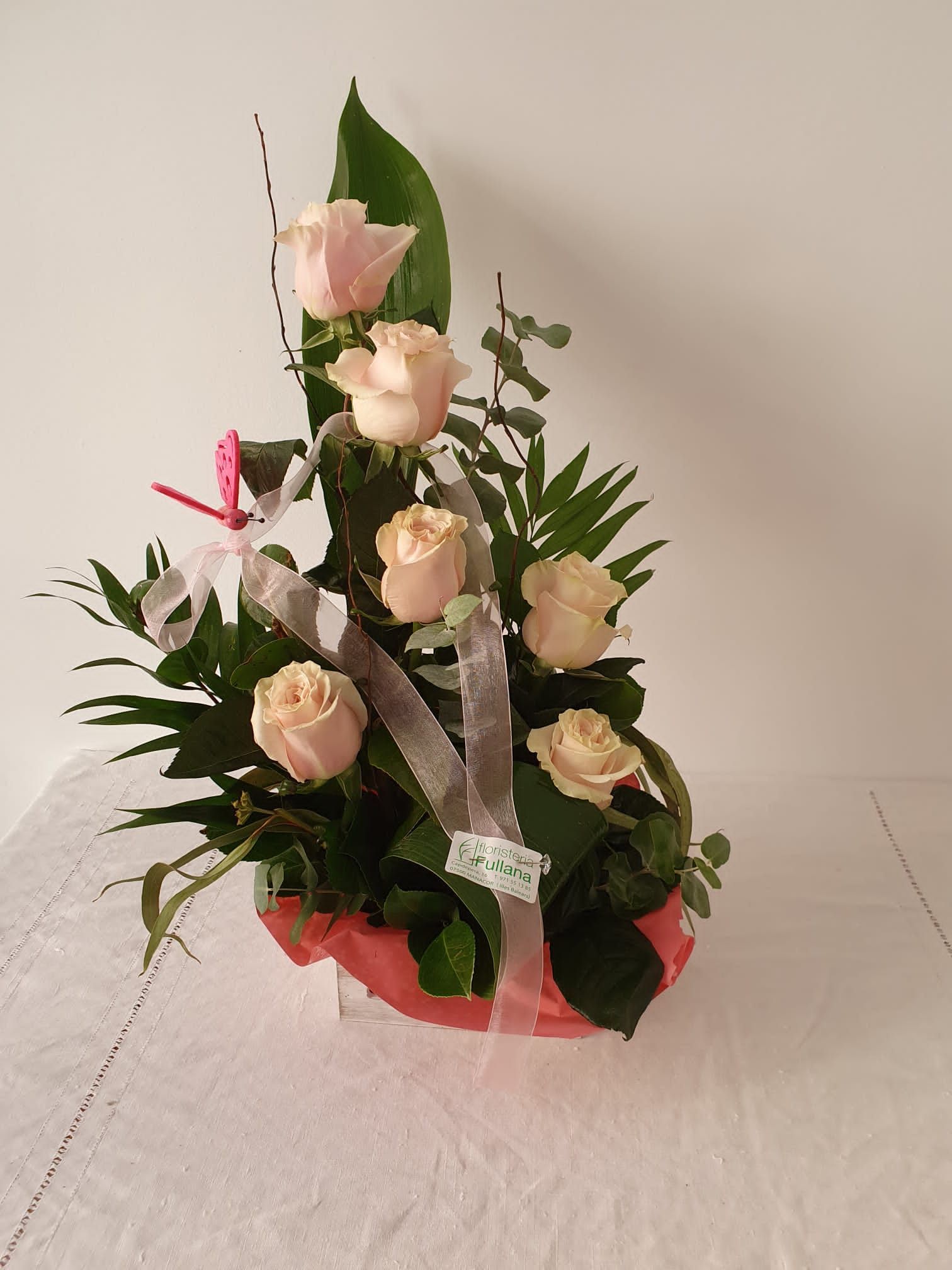 Un arreglo floral con seis rosas de color rosa claro, vegetación y cinta, presentado en una canasta roja sobre un fondo blanco.