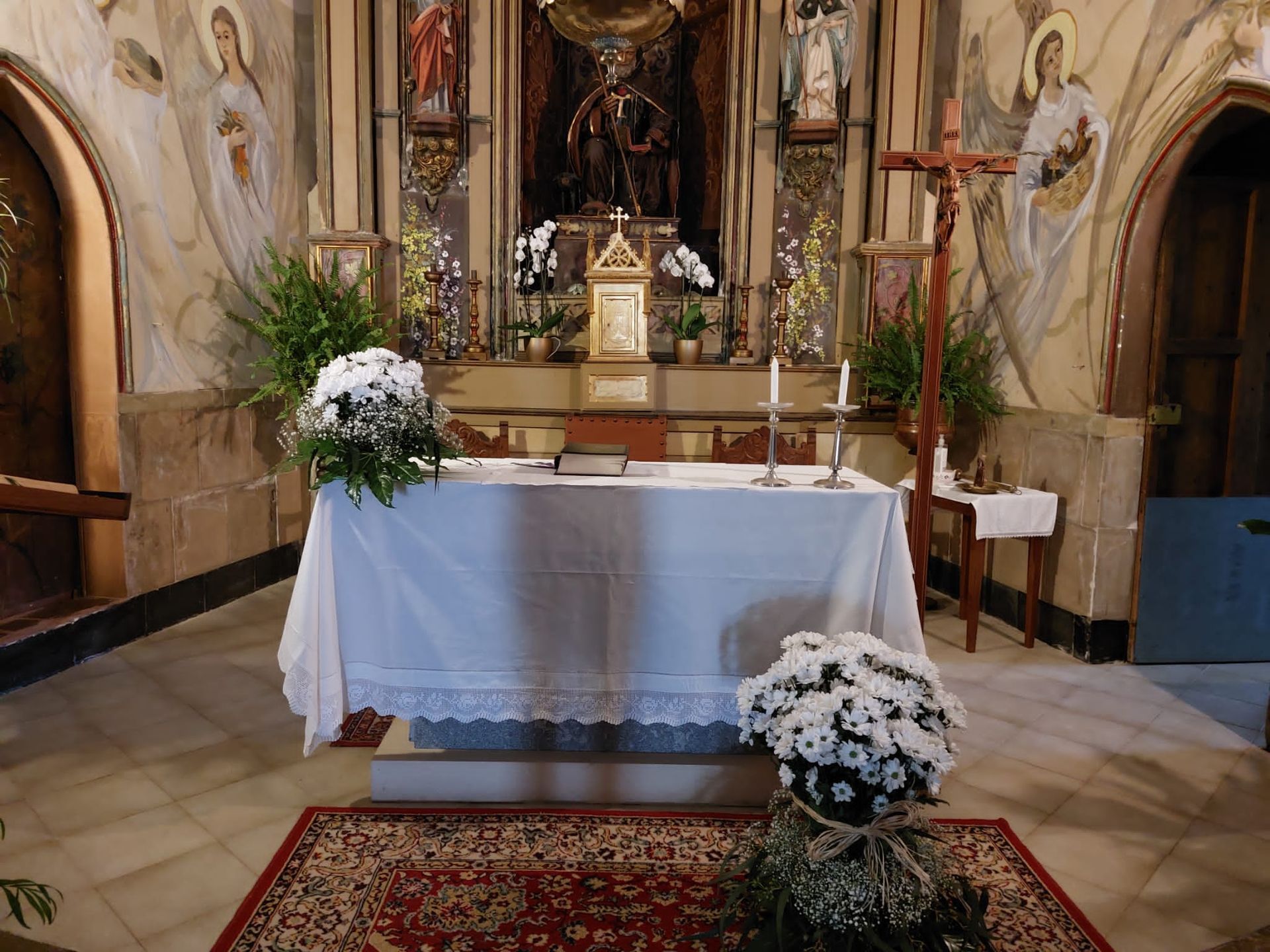 Interior de una iglesia con altar. Mantel blanco, flores, velas y un crucifijo. Paredes beige con pinturas.