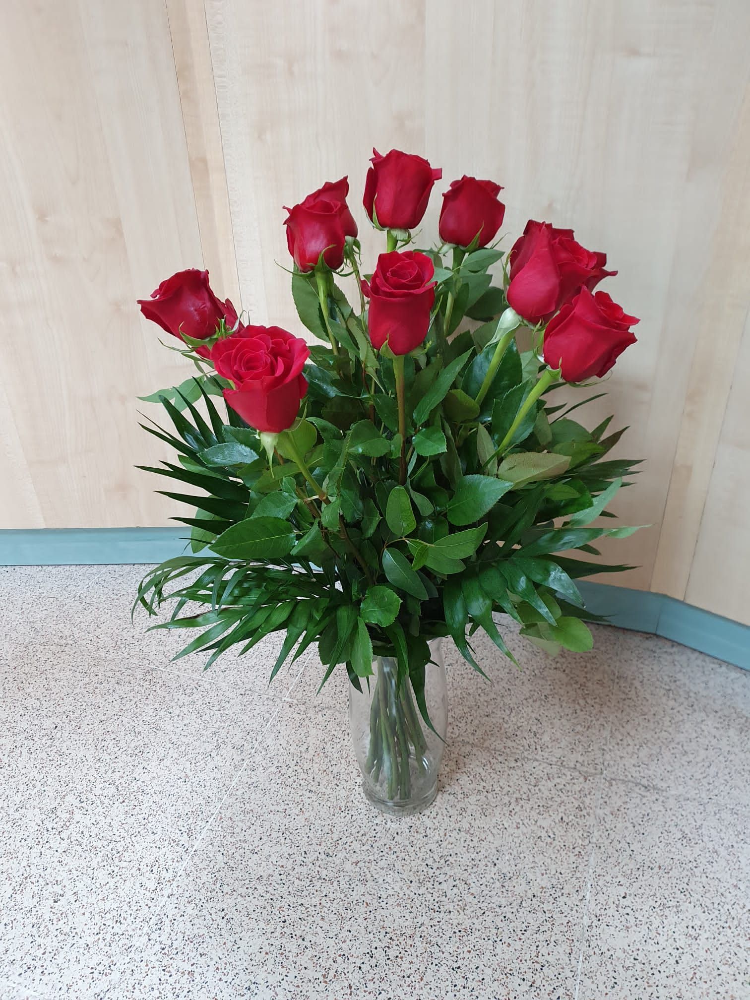 Un ramo de rosas rojas en un jarrón transparente con follaje verde, sobre un suelo de baldosas de color beige y gris.