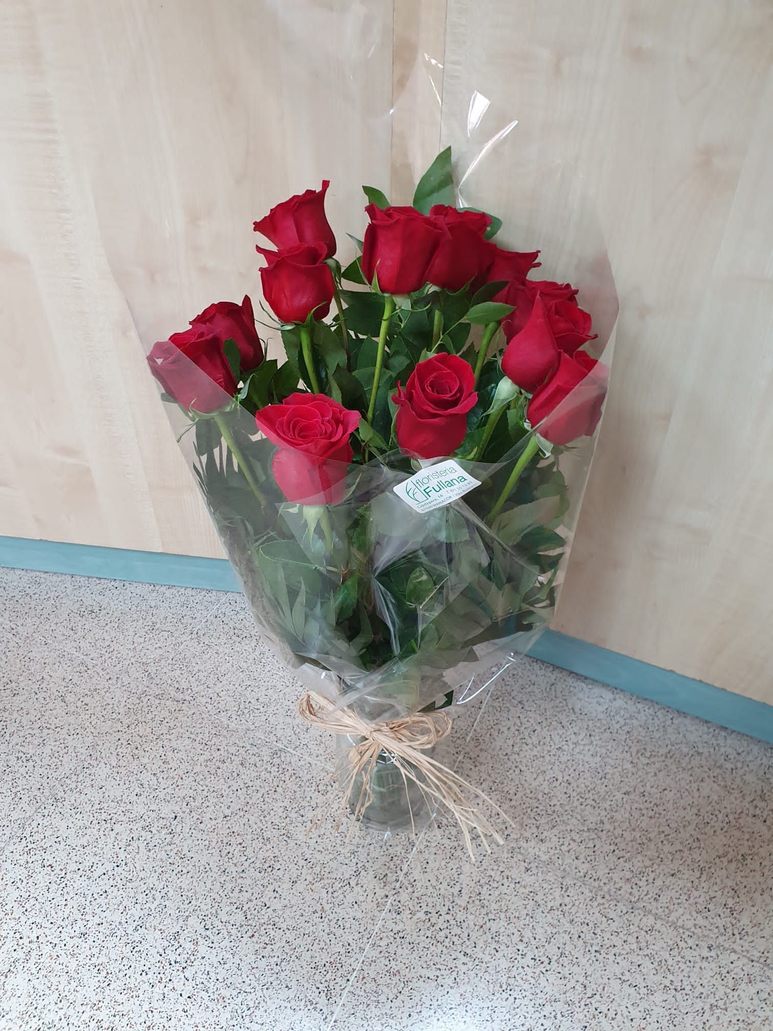 Un ramo de rosas rojas envuelto en celofán, atado con cordel, sobre un suelo de baldosas contra una pared revestida de madera.