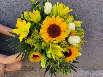 Una persona sostiene un ramo brillante con lirios amarillos, girasoles y flores blancas contra una pared gris.