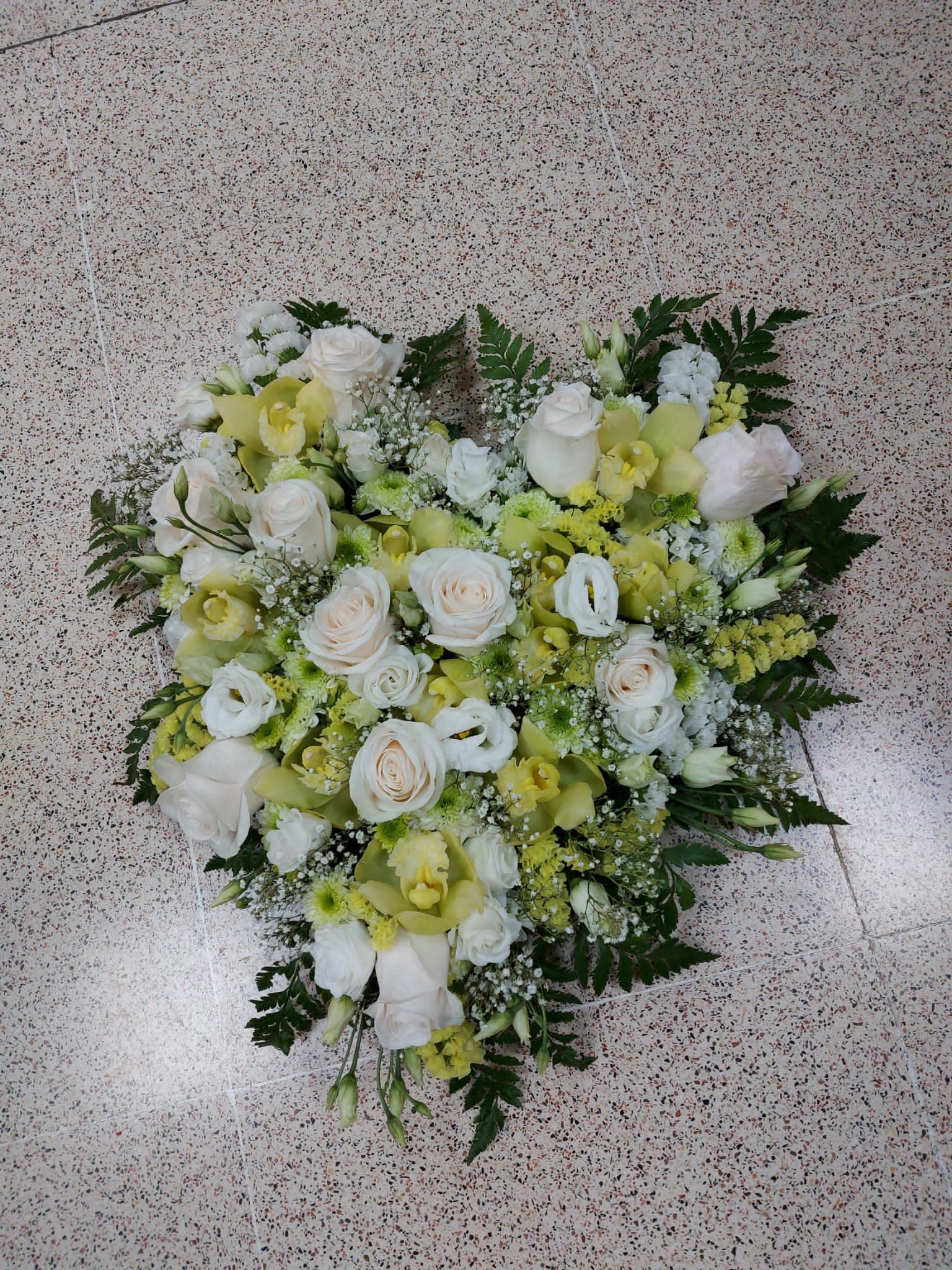Arreglo floral en forma de corazón con rosas blancas, orquídeas amarillas y vegetación, sobre un piso moteado.