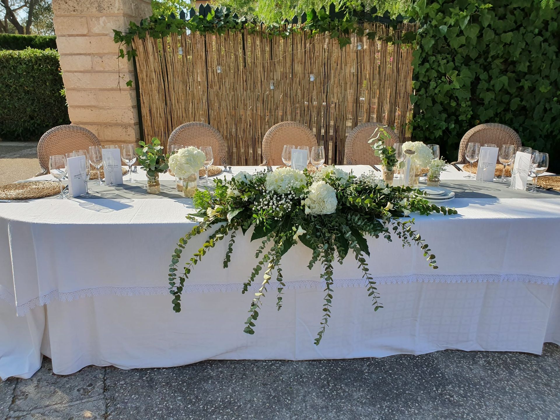 Una mesa al aire libre decorada, preparada para una reunión, con un mantel blanco, centros de mesa florales y manteles individuales tejidos.