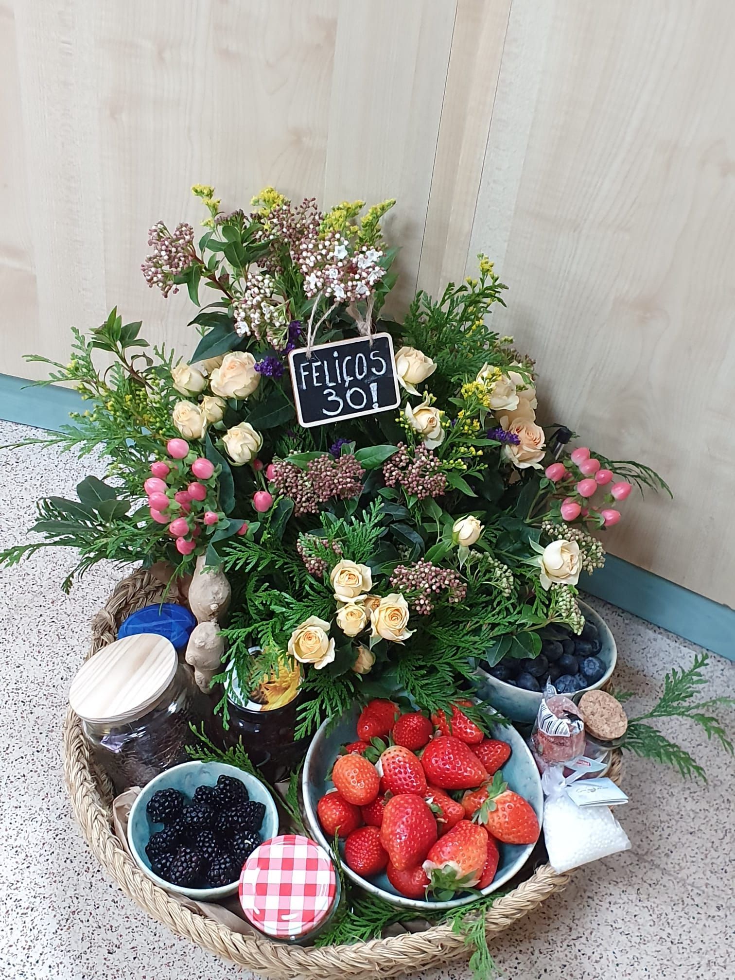 Una canasta tejida rebosante de flores, bayas y frascos de conservas, con un cartel que dice