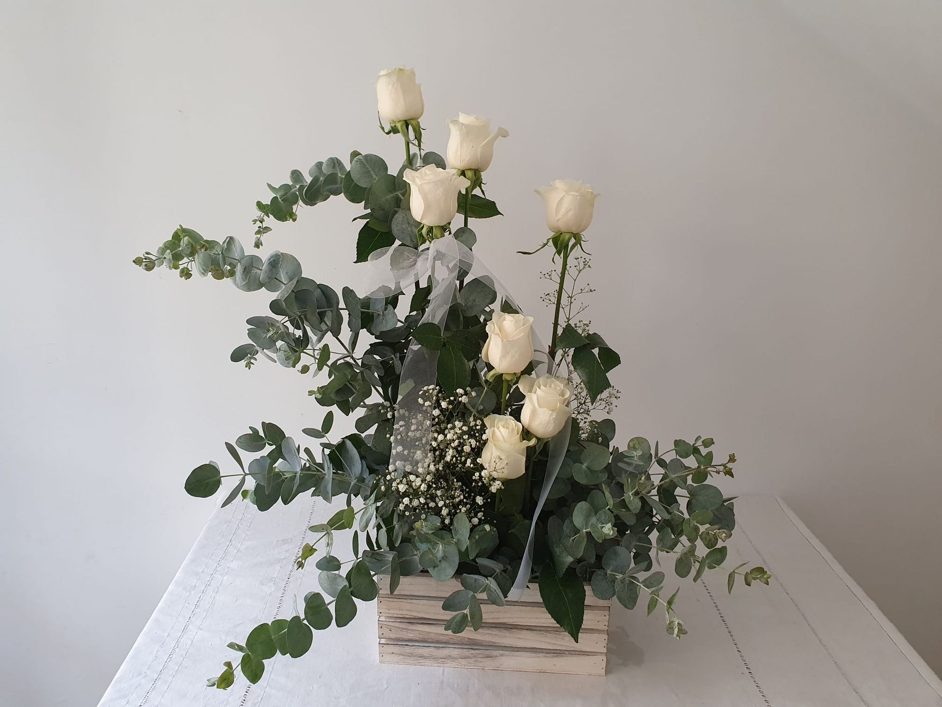 Rosas blancas en una caja de madera con hojas de eucalipto. El arreglo se presenta sobre un mantel blanco contra una pared blanca.
