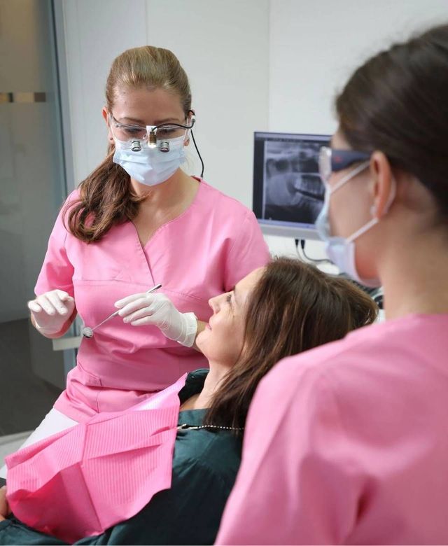 Eine Zahnärztin in rosa Uniform und mit Lupenbrille behandelt einen Patienten, unterstützt von einer weiteren Mitarbeiterin in einer Zahnarztpraxis.