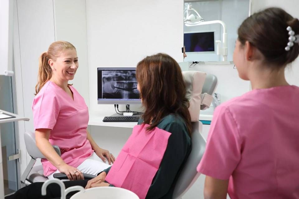 Eine professionelle Zahnärztin in rosa OP-Kleidung lächelt, während sie mit einem Patienten auf dem Zahnarztstuhl Bilder der Zahnvorsorge auf einem Monitor bespricht.