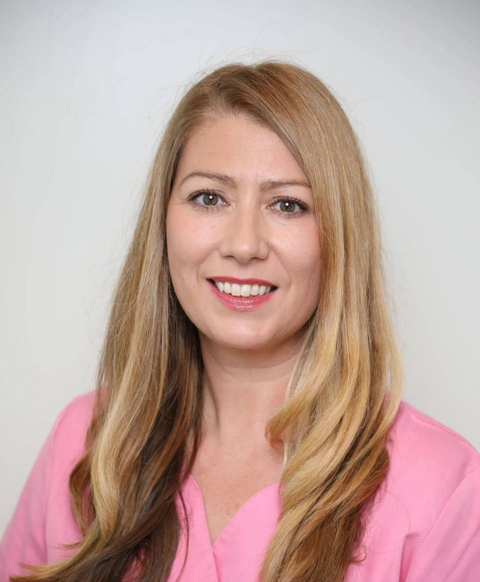 Eine Person mit langen, blond gesträhnten Haaren, die ein rosa V-Ausschnitt-Top trägt und vor einem schlichten weissen Hintergrund lächelt.