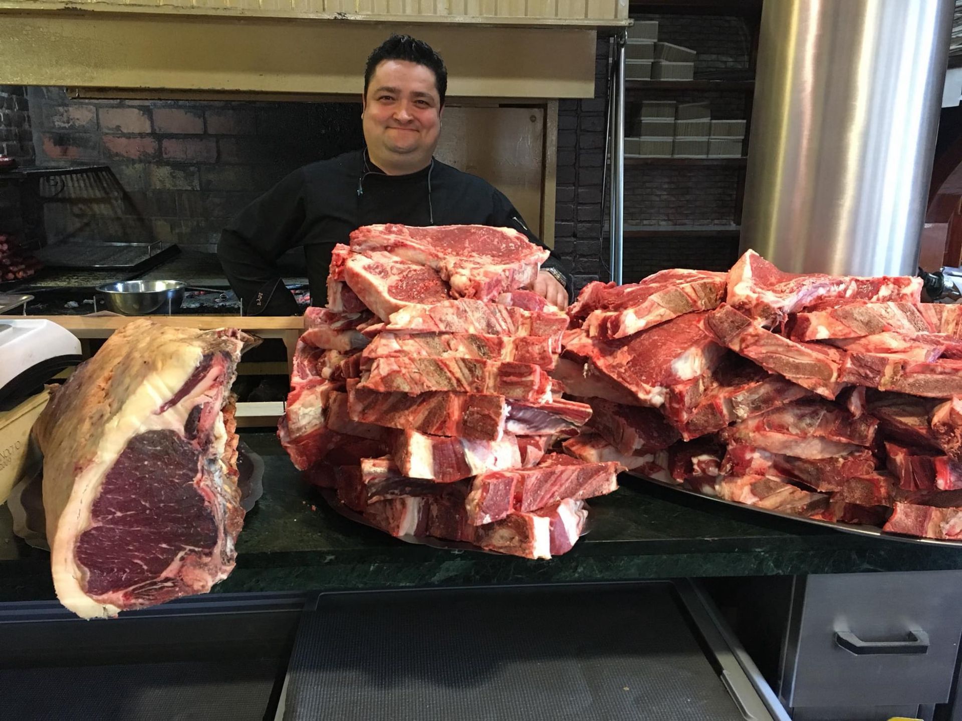 Chef detrás de un mostrador con una gran exhibición de filetes crudos y cortados, con un horno abierto al fondo.