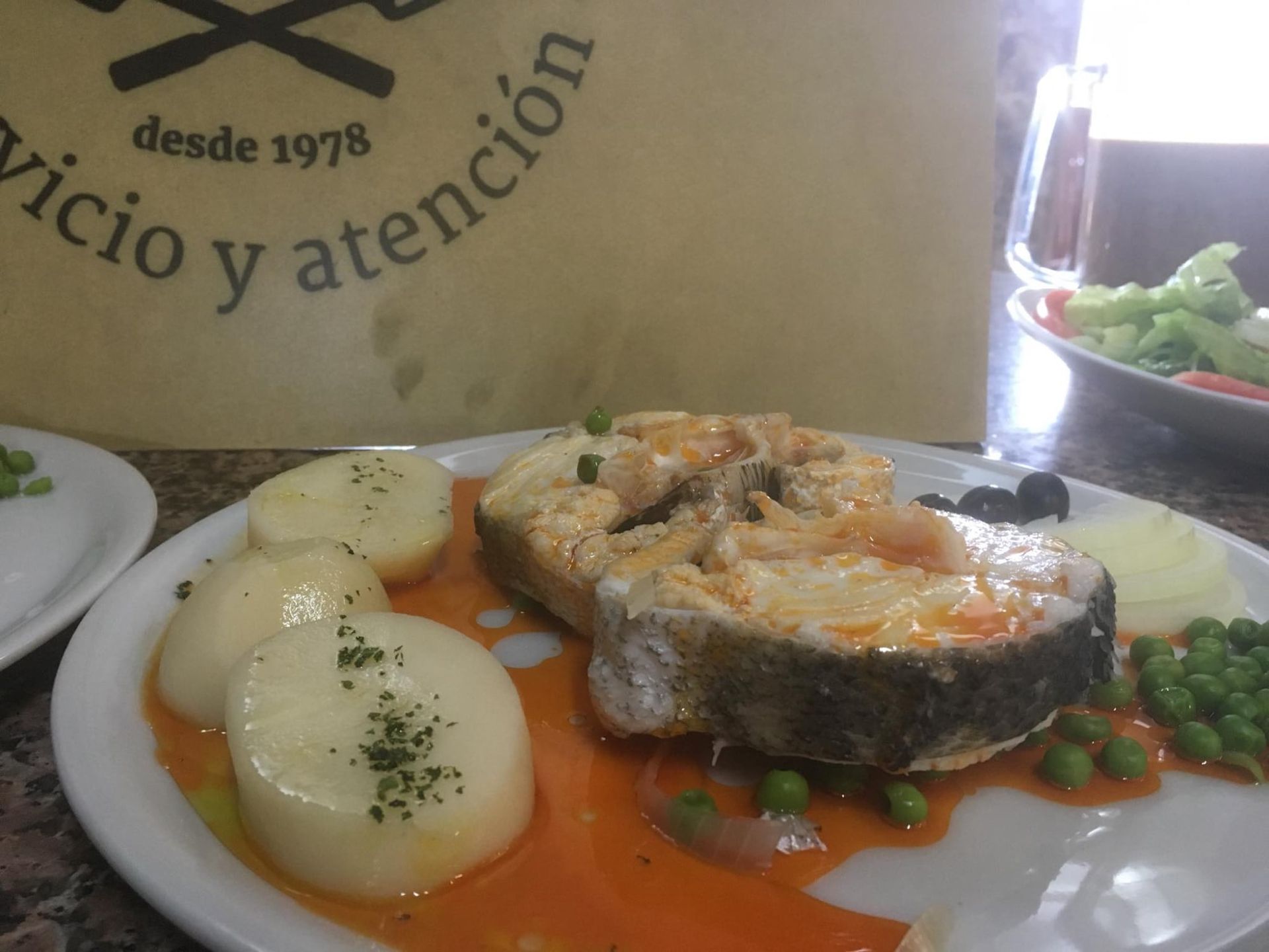 Plato con pescado, patatas, guisantes y salsa. En un restaurante, junto a un plato de guisantes y ensalada.