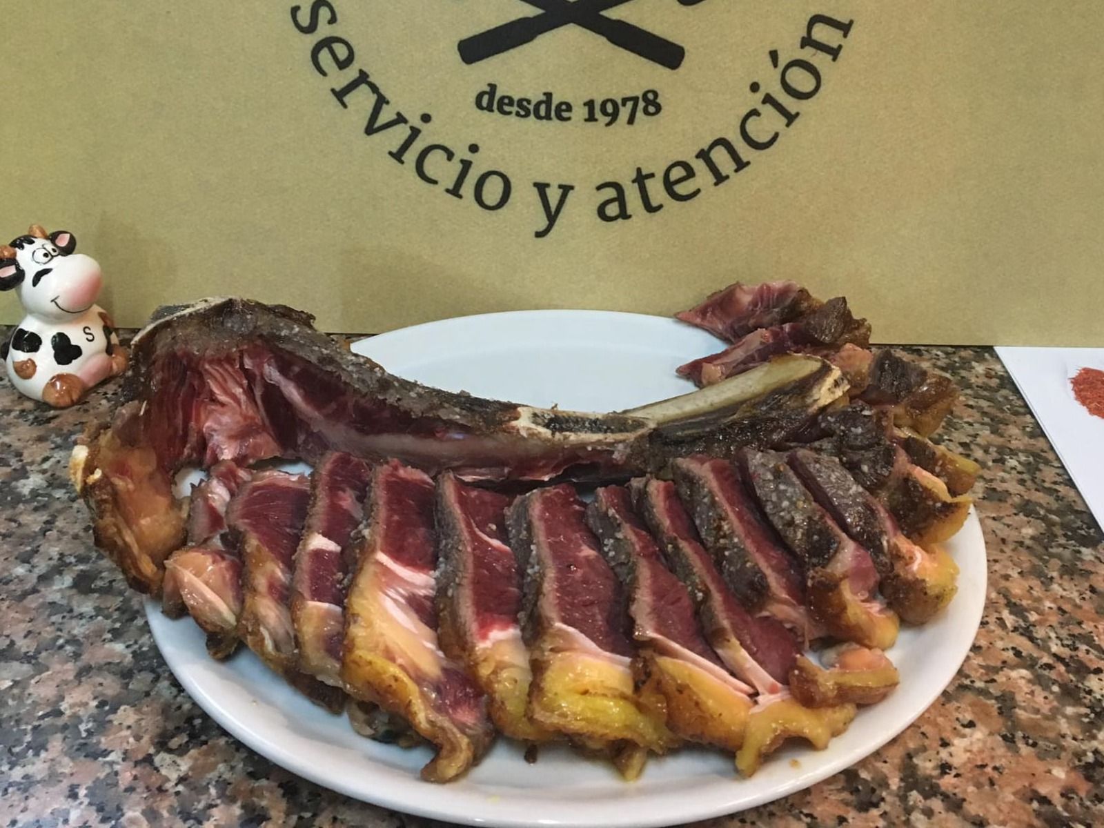 Filete rebanado en un plato; una figura de vaca a la izquierda. Letrero de restaurante al fondo.