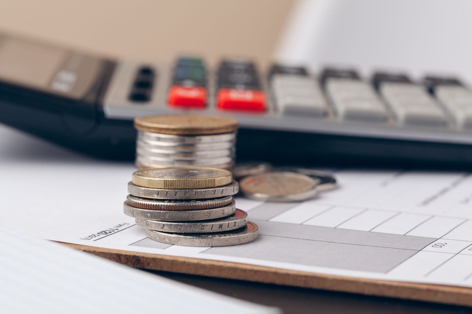 Monedas apiladas en un portapapeles junto a una calculadora y un bolígrafo sobre un escritorio.