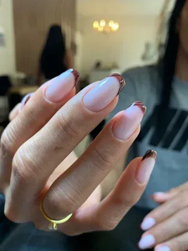 Primer plano de una mano con uñas pintadas de color rosa pálido y anillos de oro, sostenida frente a una persona borrosa en un espacio interior.