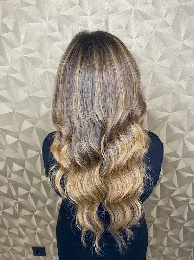 Cabello largo y ondulado rubio con mechas balayage visto desde atrás contra una pared texturizada de color beige.