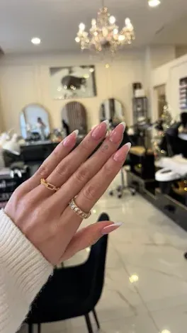 Una mano con uñas rosas y anillos dorados se alza en un elegante salón con espejos y lámparas de araña.