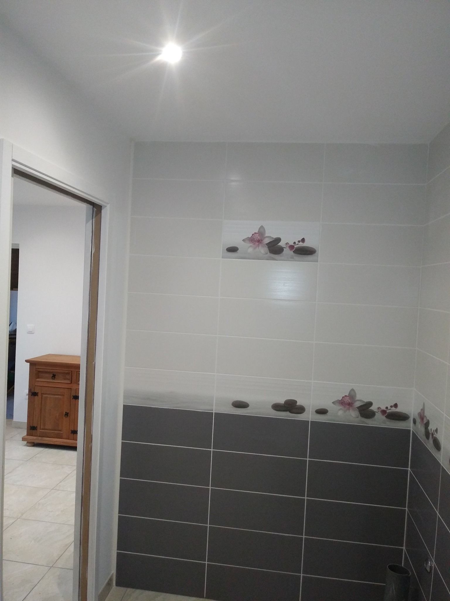 Un couloir avec des carreaux blancs et gris, un luminaire et une vue sur une autre pièce avec une armoire en bois.