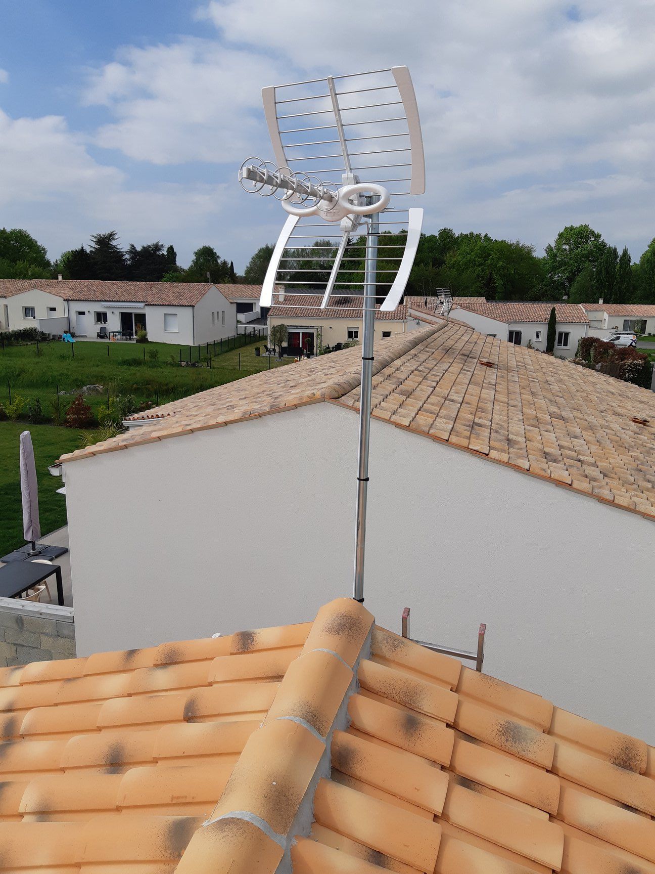 Toit avec panneaux solaires et antenne, avec maisons et arbres en arrière-plan sous un ciel bleu.