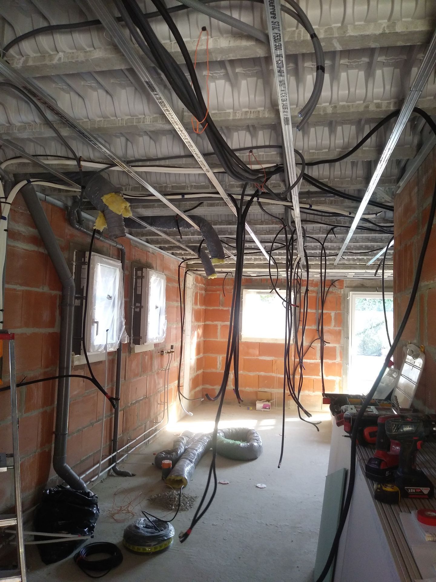Chantier intérieur : câblage électrique apparent et supports métalliques au plafond, murs en briques, fenêtres.