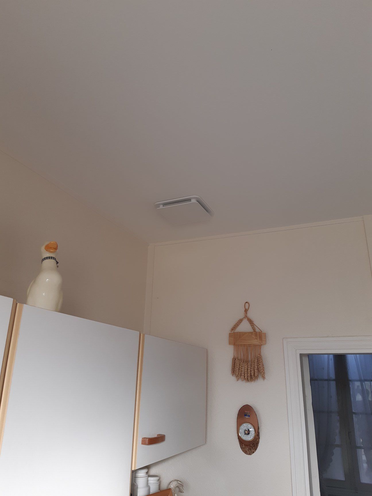 Intérieur de cuisine avec armoires blanches, ventilation au plafond et objets décoratifs sur le mur et le dessus des armoires.