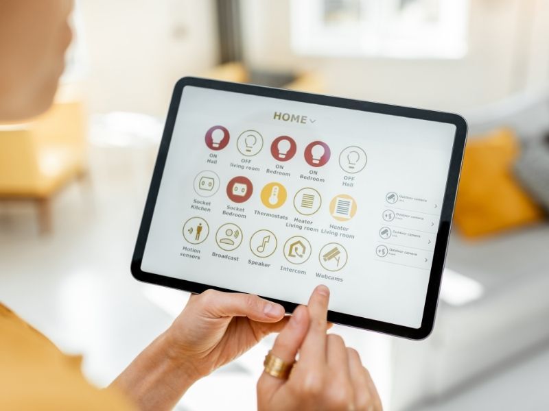 Une personne utilise une tablette pour contrôler ses appareils domestiques connectés. L'écran affiche des icônes pour les lumières, les portes et la climatisation.