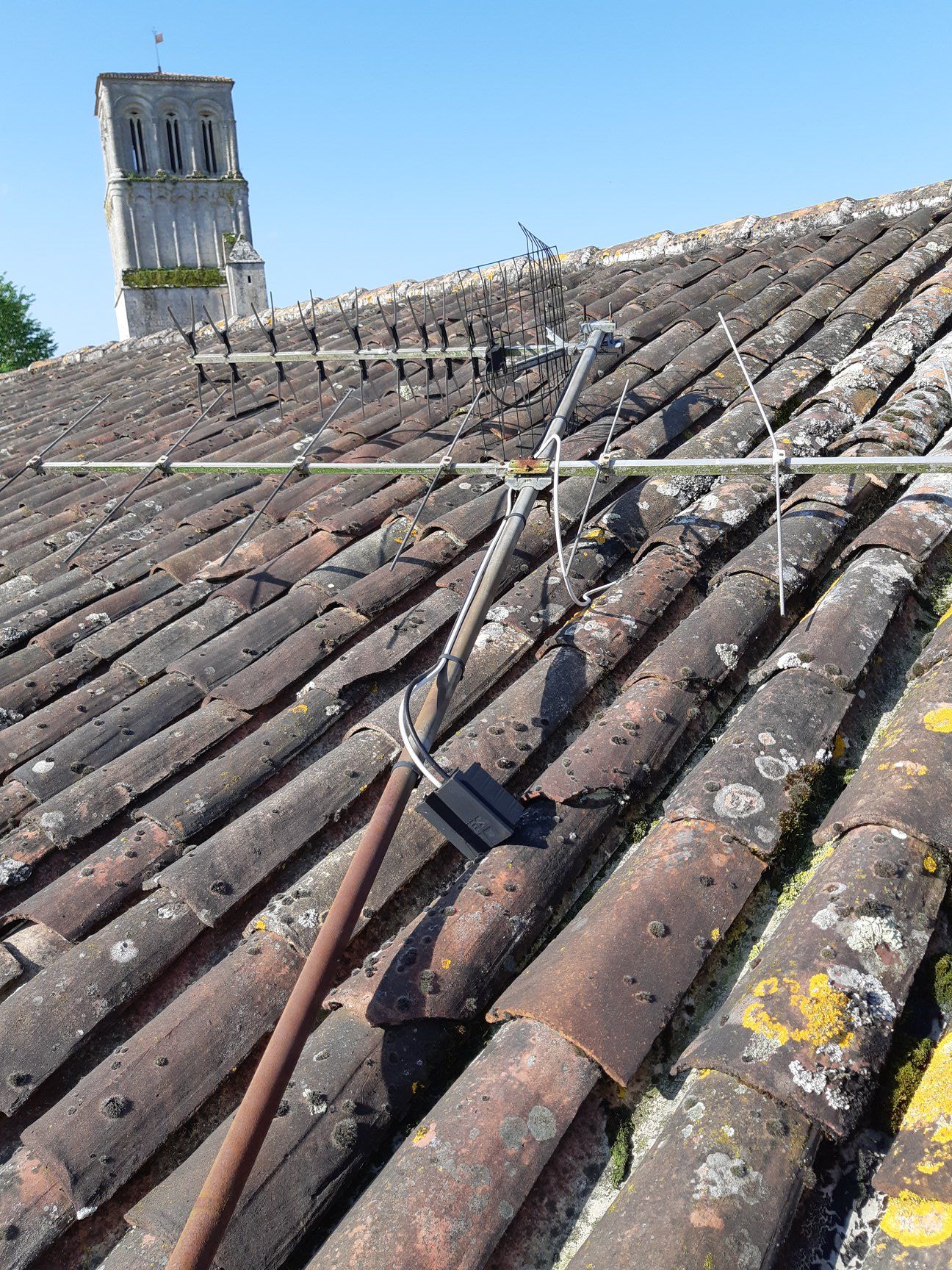 Une antenne rouillée sur un toit de tuiles, avec un clocher d'église en arrière-plan sous un ciel bleu.