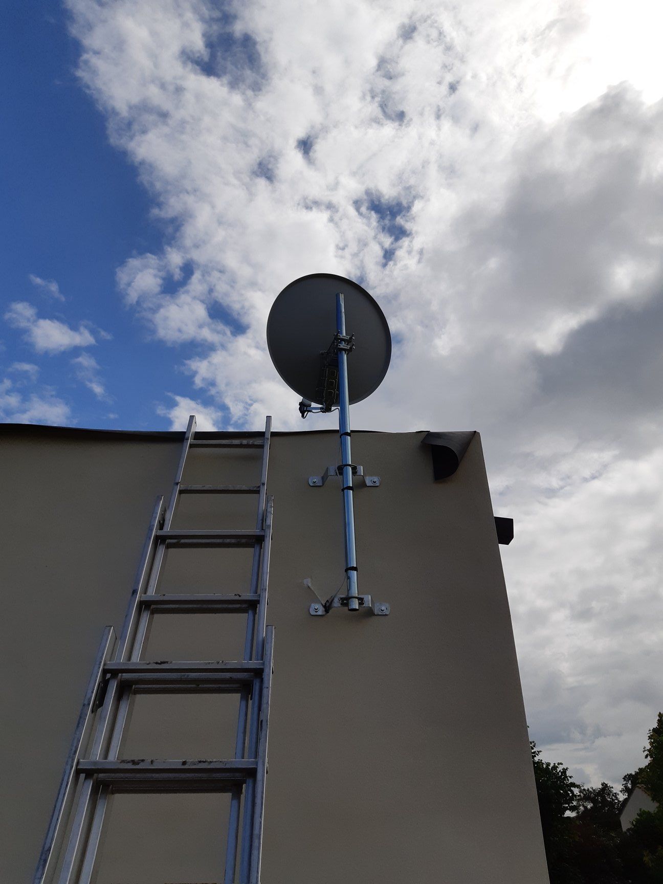 Une antenne parabolique montée sur un mur avec une échelle appuyée contre elle, fond de ciel bleu.