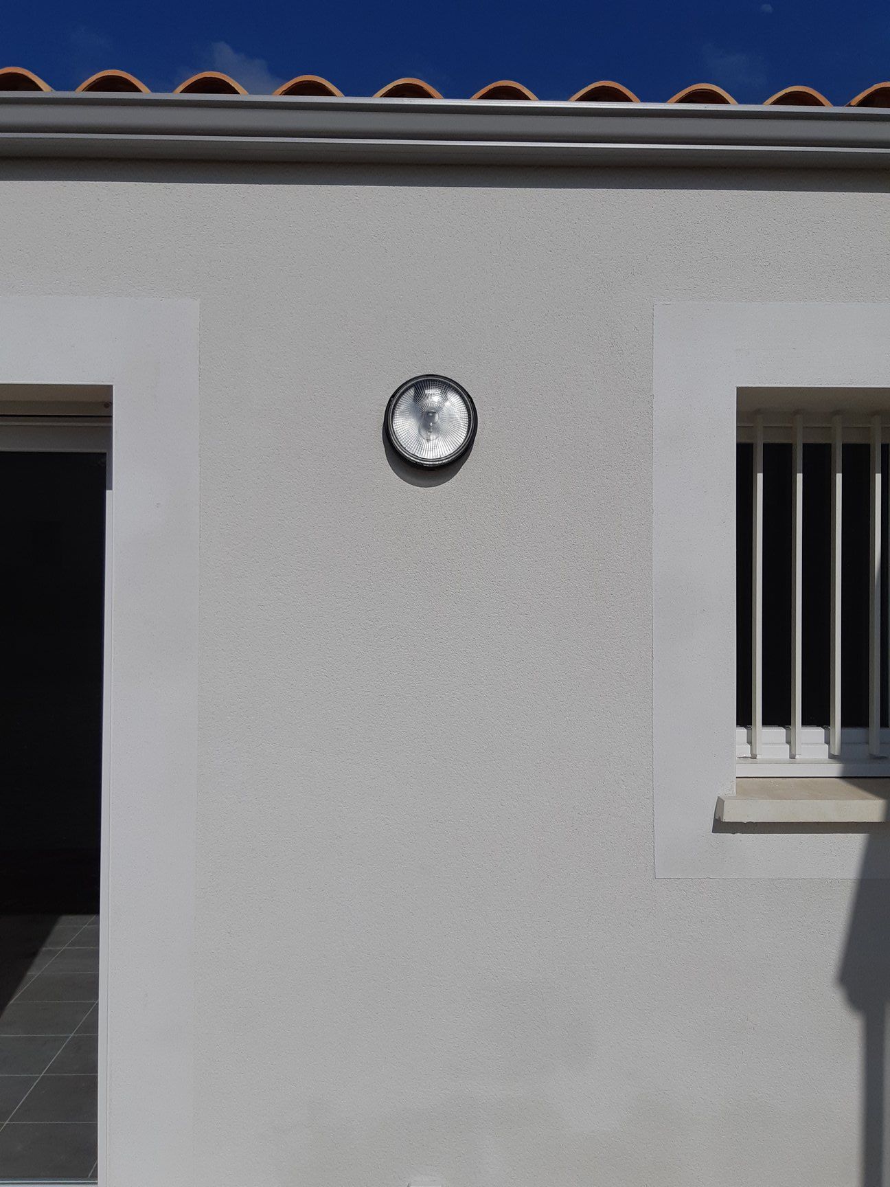 Bâtiment blanc avec un luminaire, une fenêtre avec des barreaux et une porte sous un ciel bleu.