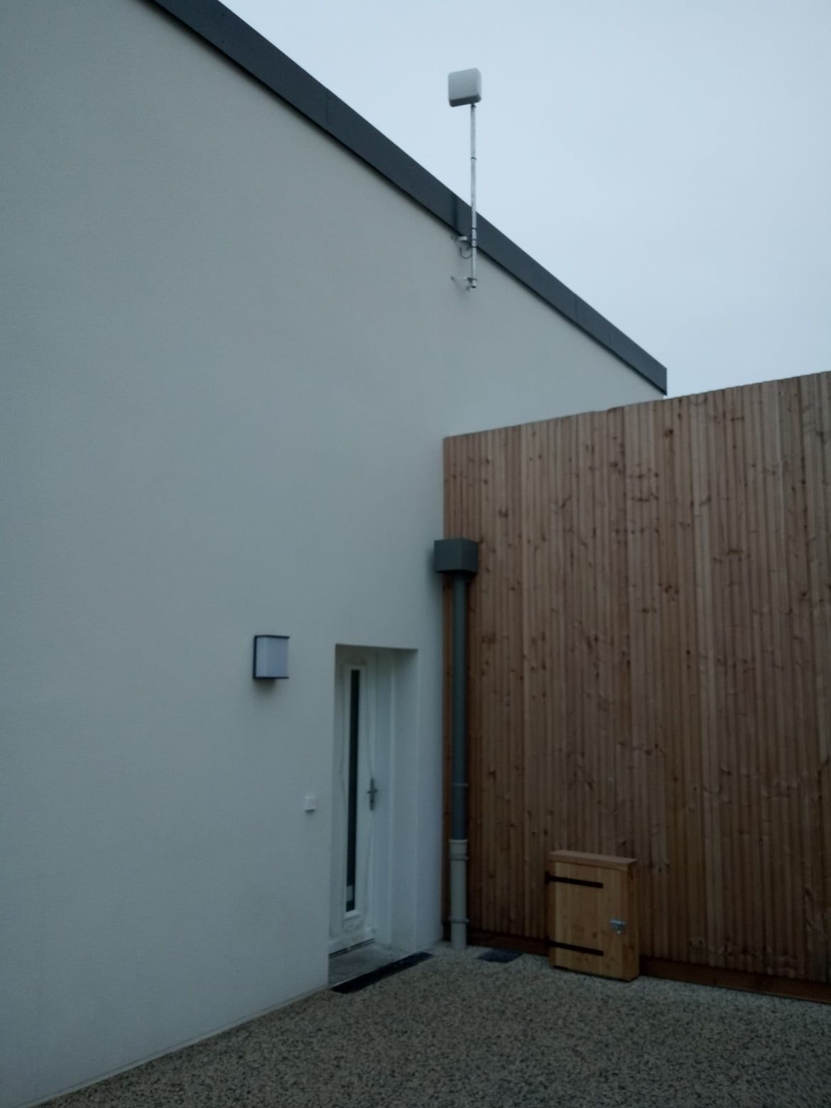 Bâtiment blanc avec une porte blanche, une clôture en bois et une zone de gravier ; une antenne est montée au-dessus.