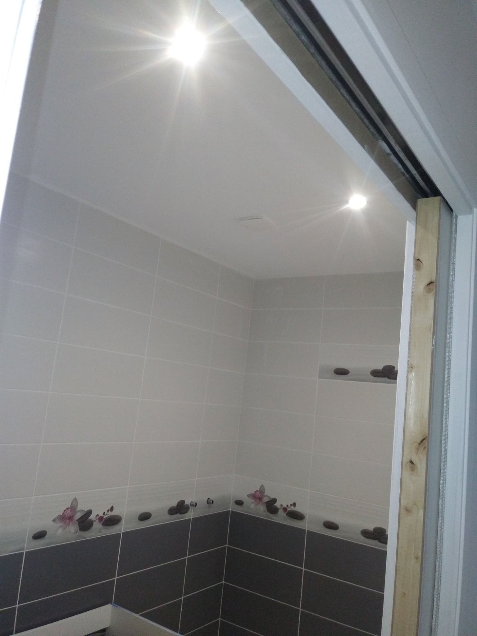 Salle de bains avec murs carrelés gris et gris foncé, plafonniers lumineux, vue à travers la porte.