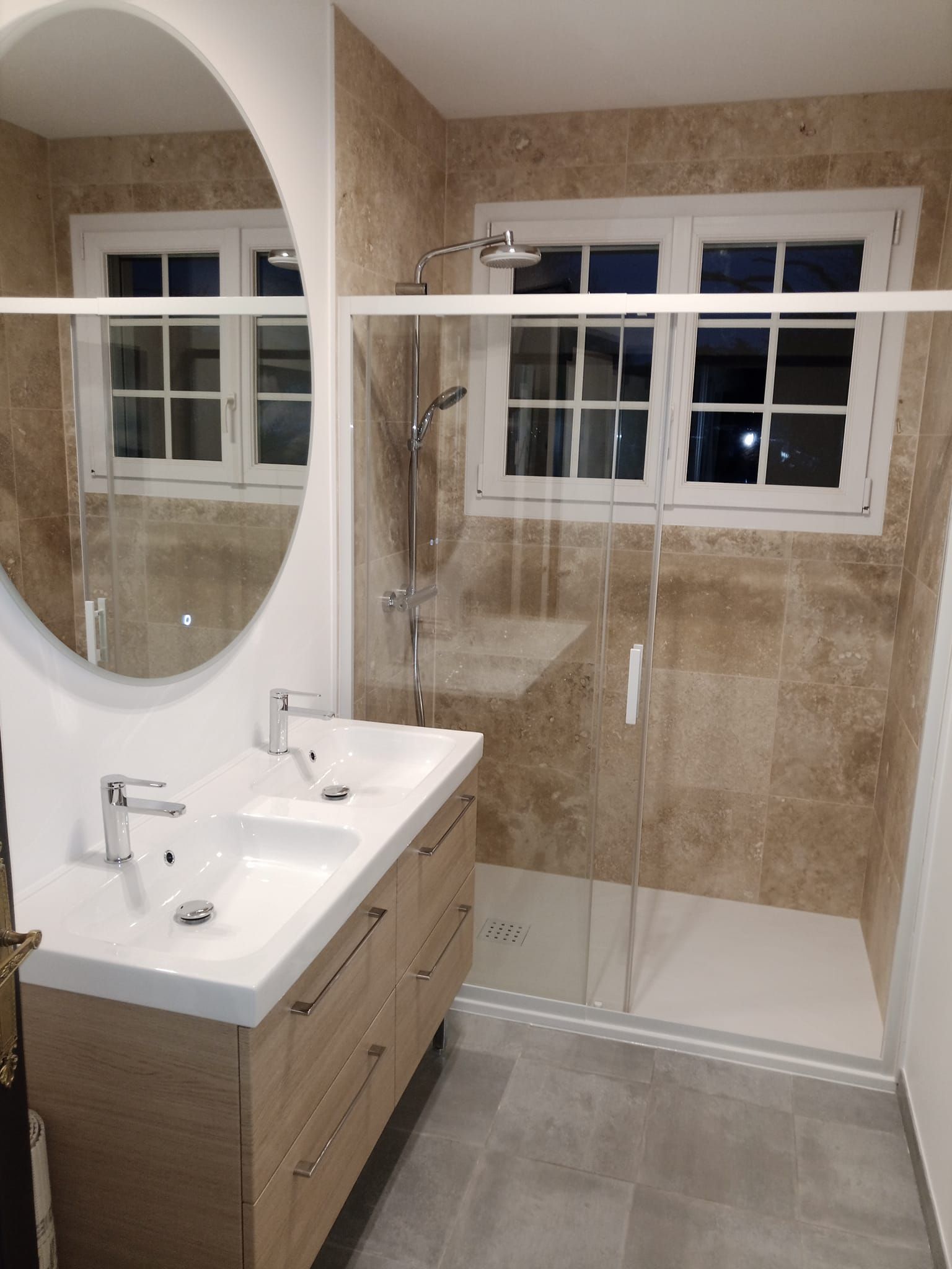Salle de bains avec double vasque, douche et miroir ovale. Murs carrelés beiges, vasque en chêne et deux fenêtres.