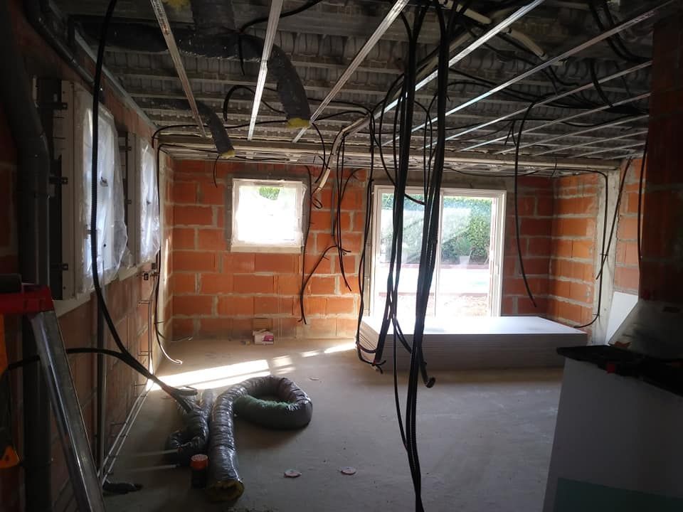 Intérieur d'une pièce en construction : murs en briques, câblage apparent, grille de plafond, fenêtres non finies et un rouleau de tube.