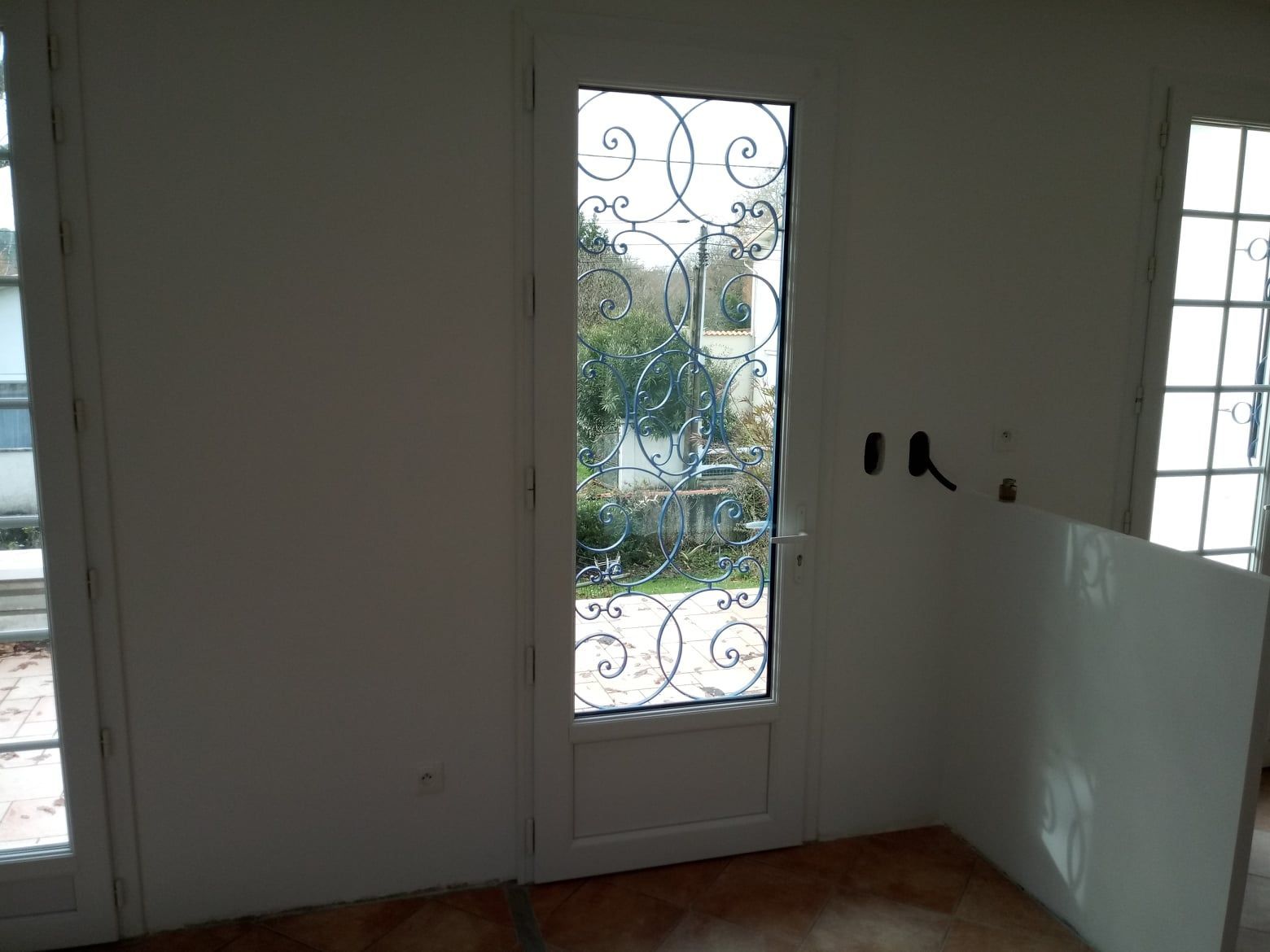 Vue intérieure d'une porte blanche avec ferronnerie décorative, menant à une vue extérieure.