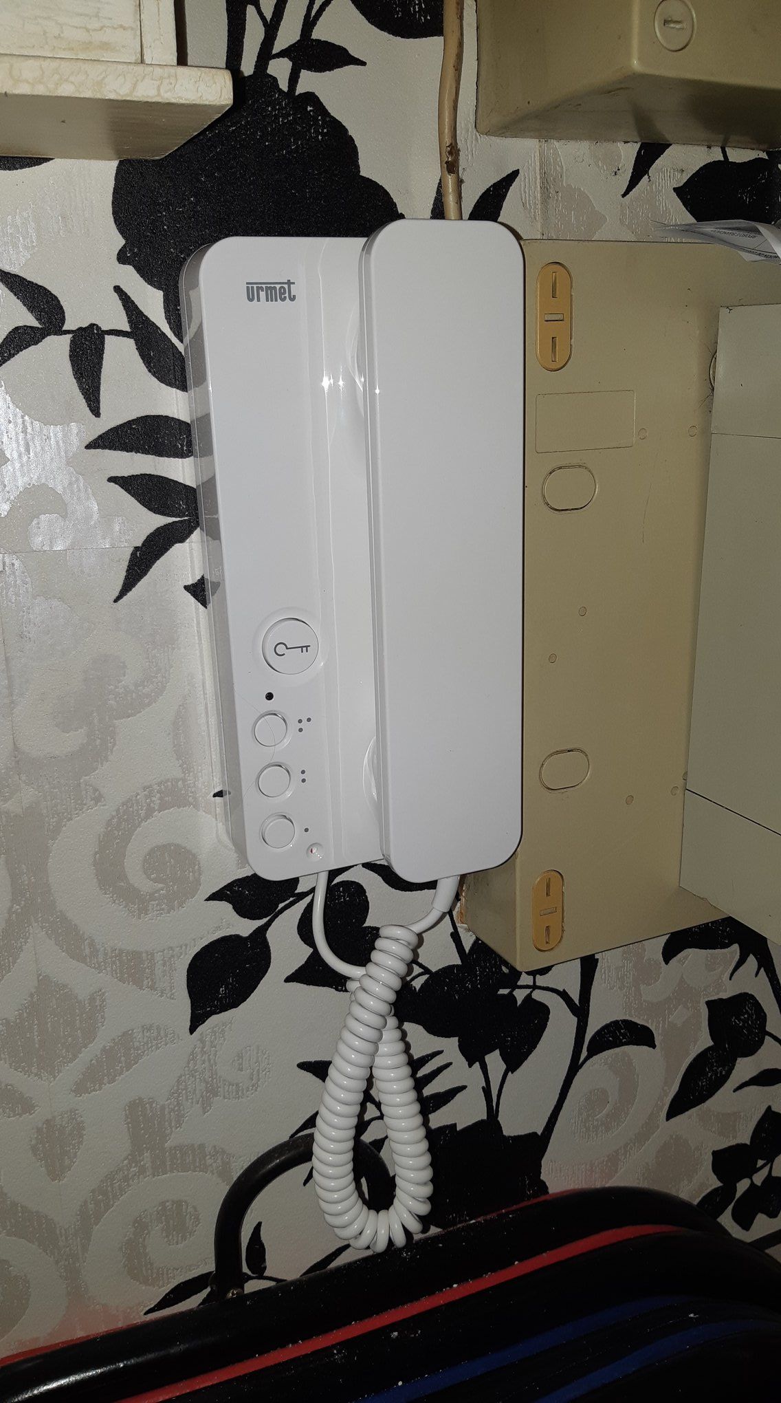 Interphone blanc sur papier peint fleuri avec lambris beige.
