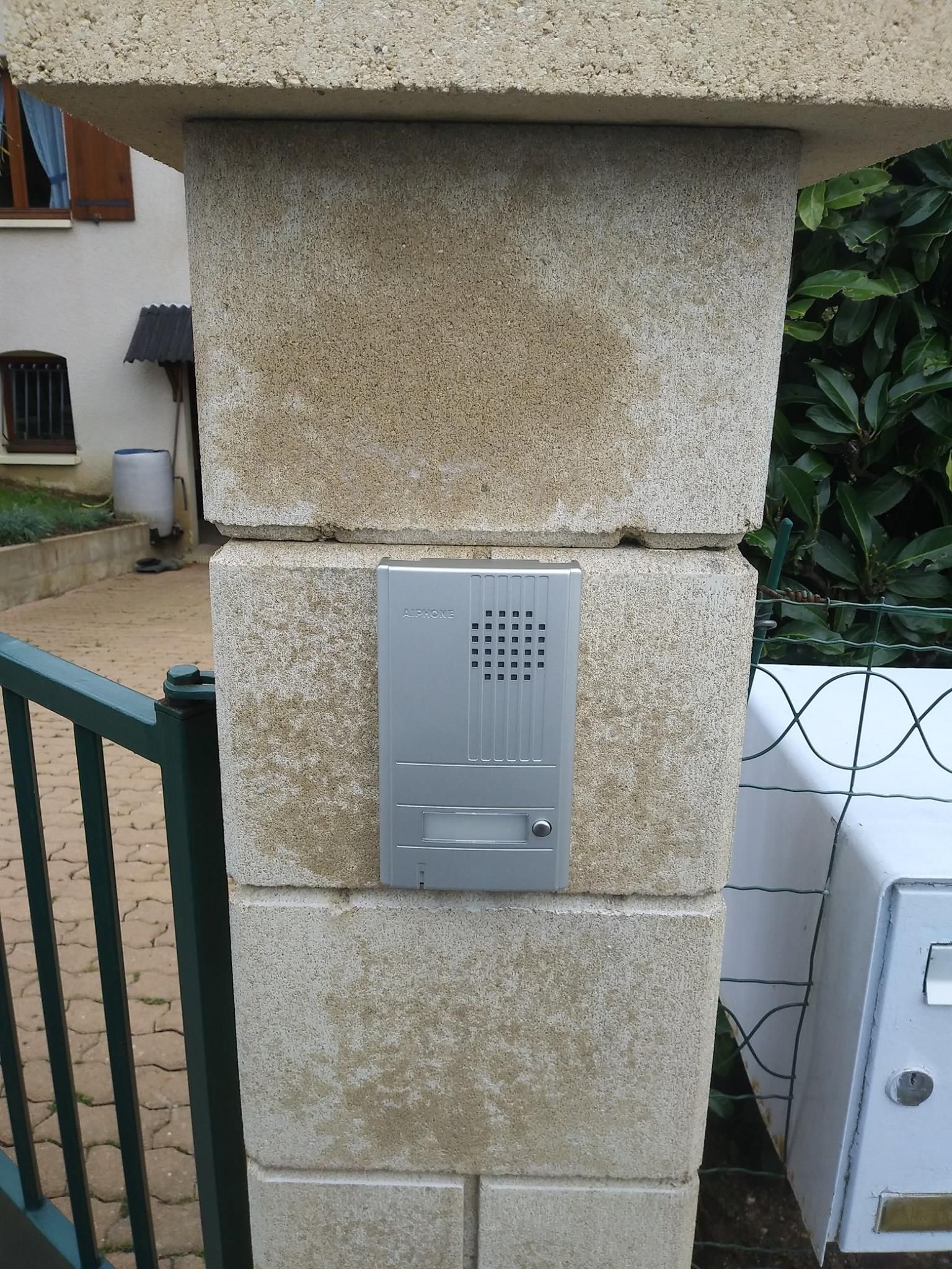 Interphone gris sur un pilier en pierre à côté d'un portail vert.