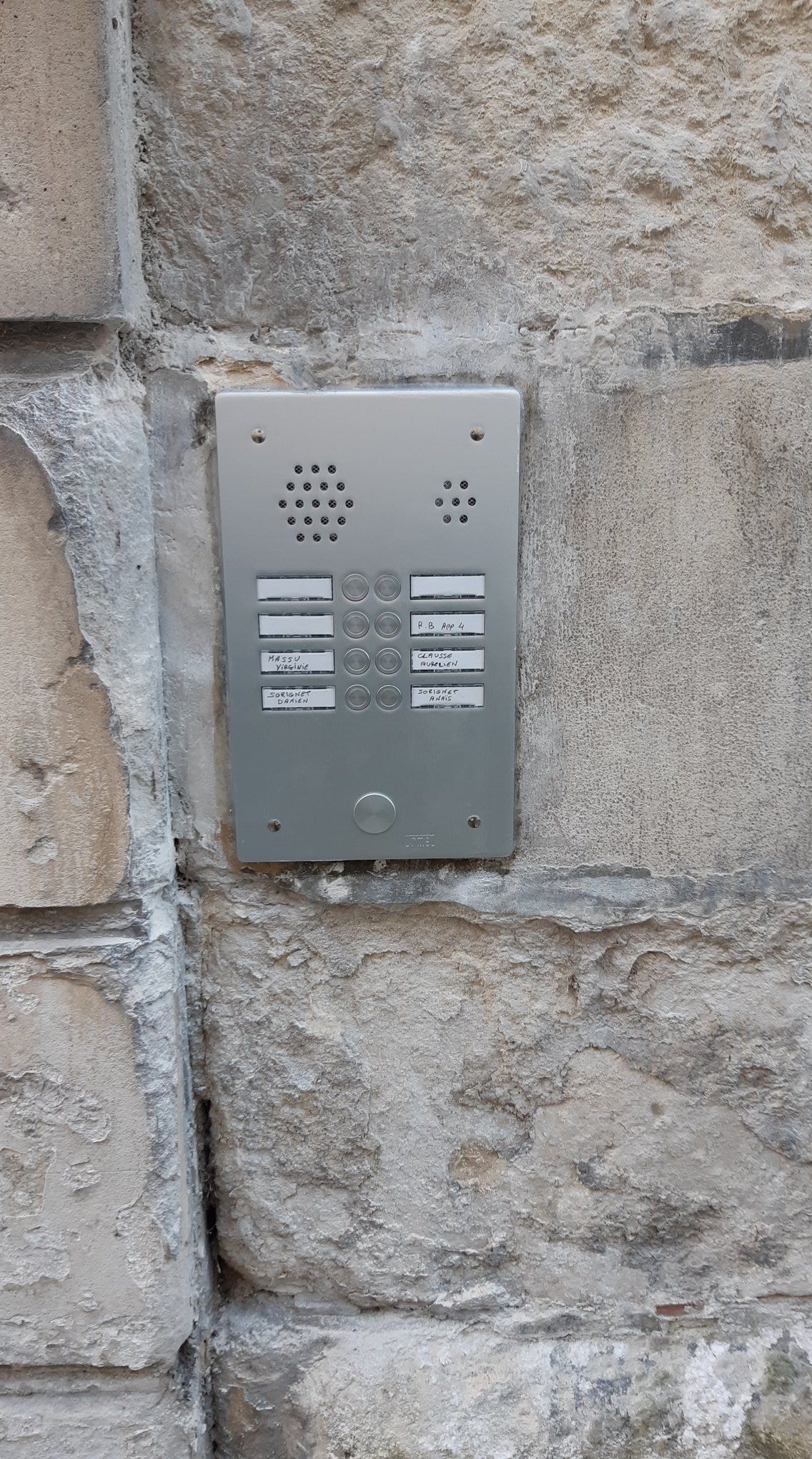 Interphone argenté sur un mur en pierre texturée avec boutons et haut-parleur.