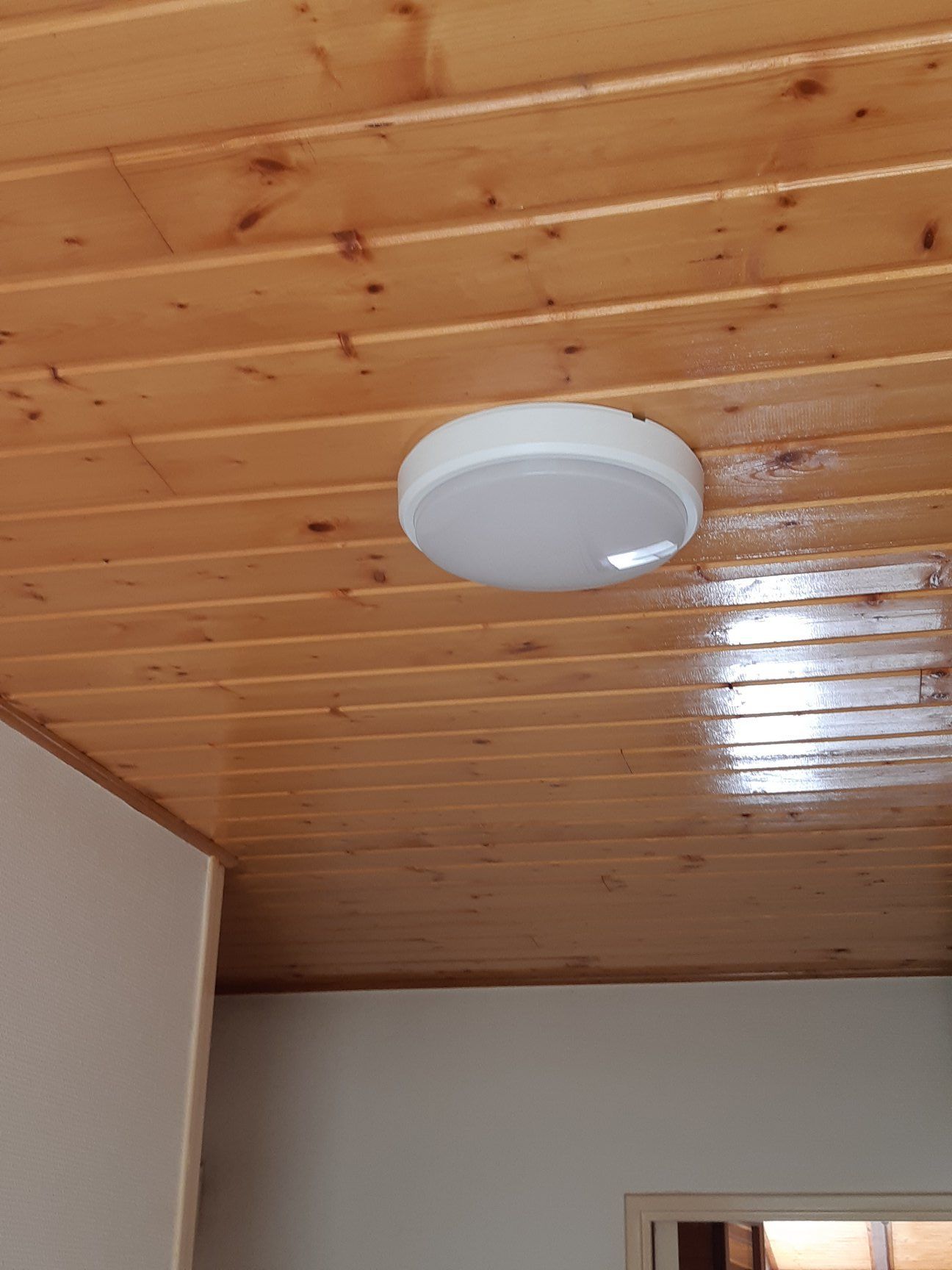 Plafonnier rond blanc sur un plafond en planches de bois teinté ; un coin d'un mur blanc est visible.