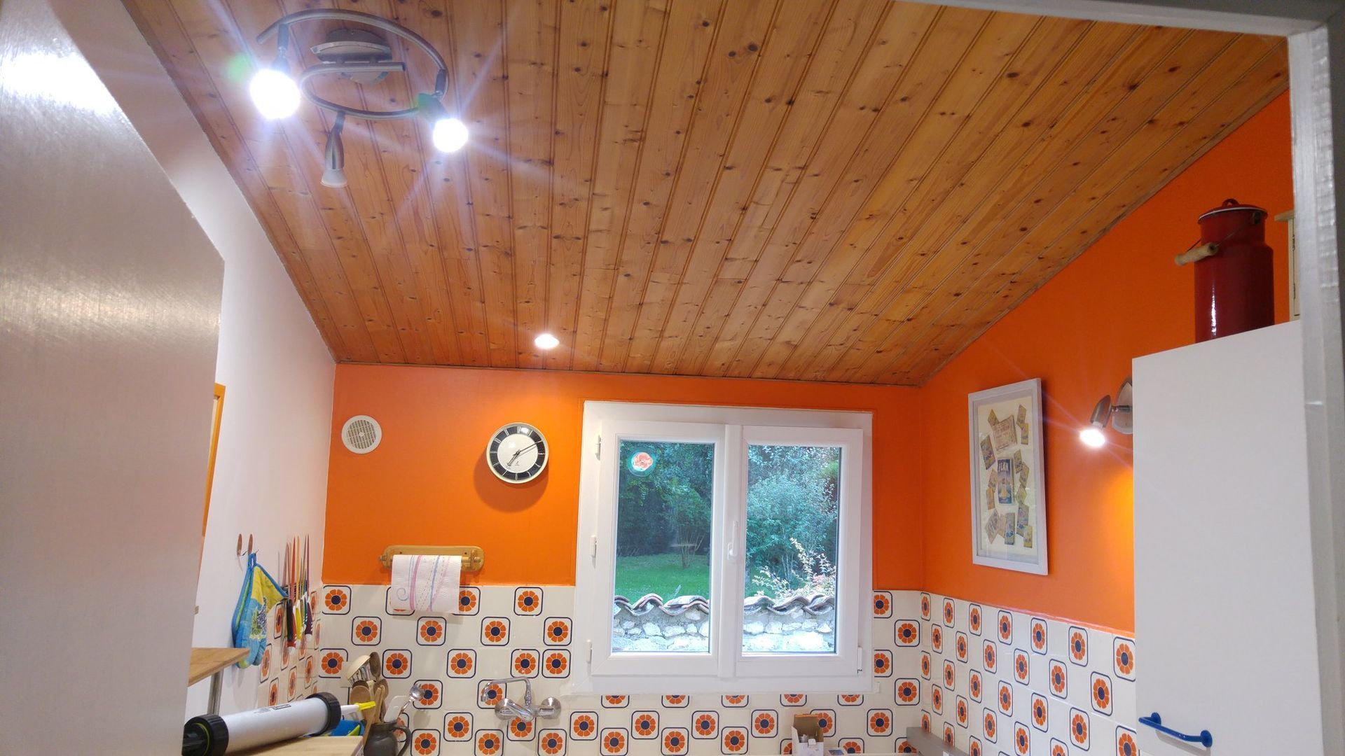Cuisine aux murs orange et au plafond lambrissé. Fenêtre, horloge et plafonniers sont visibles.