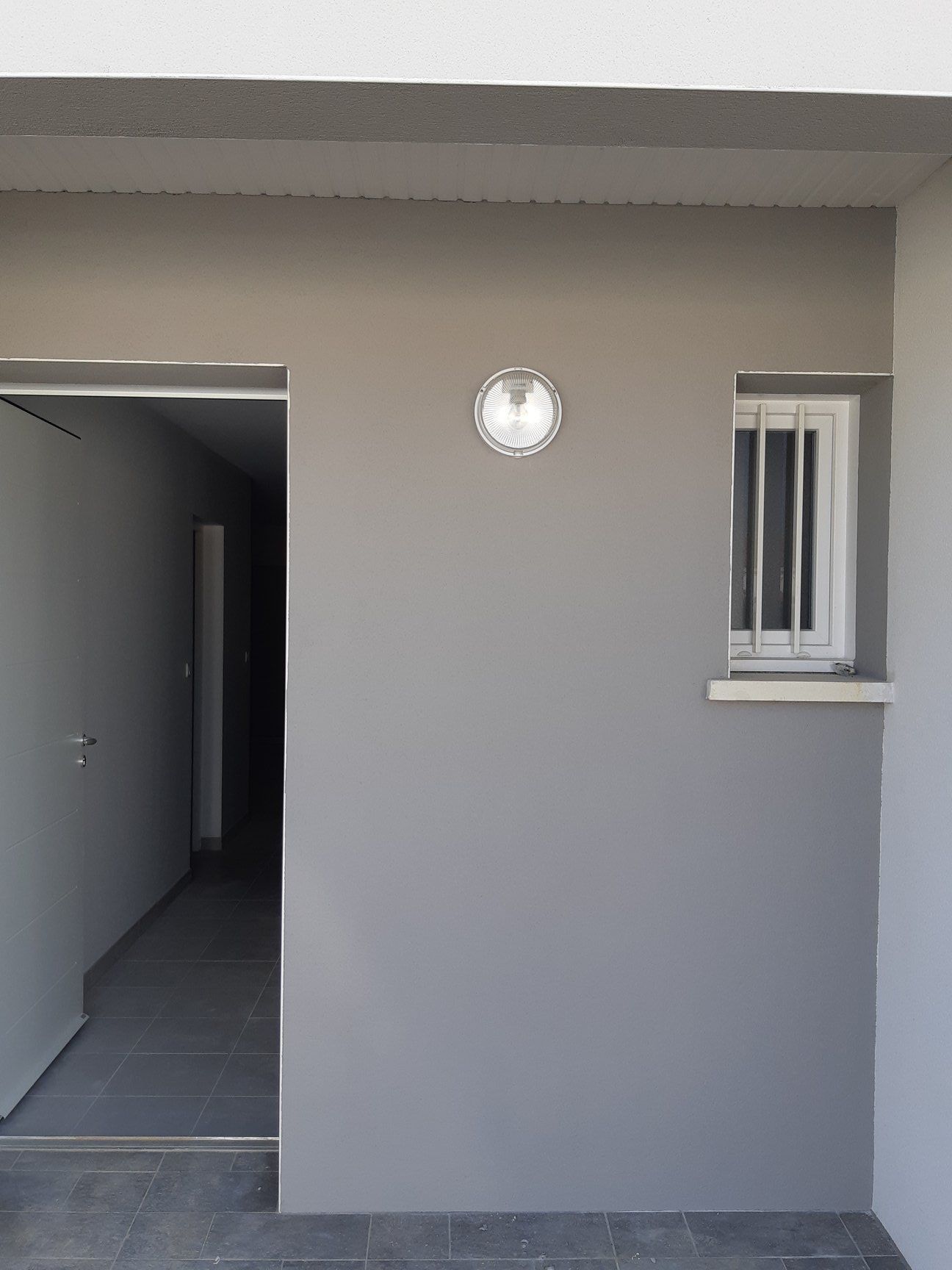 Mur extérieur gris avec porte, luminaire rond et petite fenêtre à barreaux.