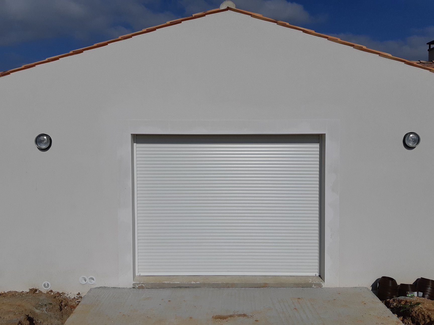 Garage blanc avec porte à enroulement fermée ; deux luminaires muraux, rampe en béton.
