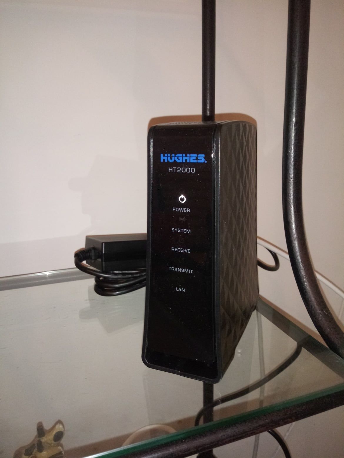 Modem Internet par satellite noir Hughes HT2000 sur une étagère en verre, avec son adaptateur secteur à proximité.