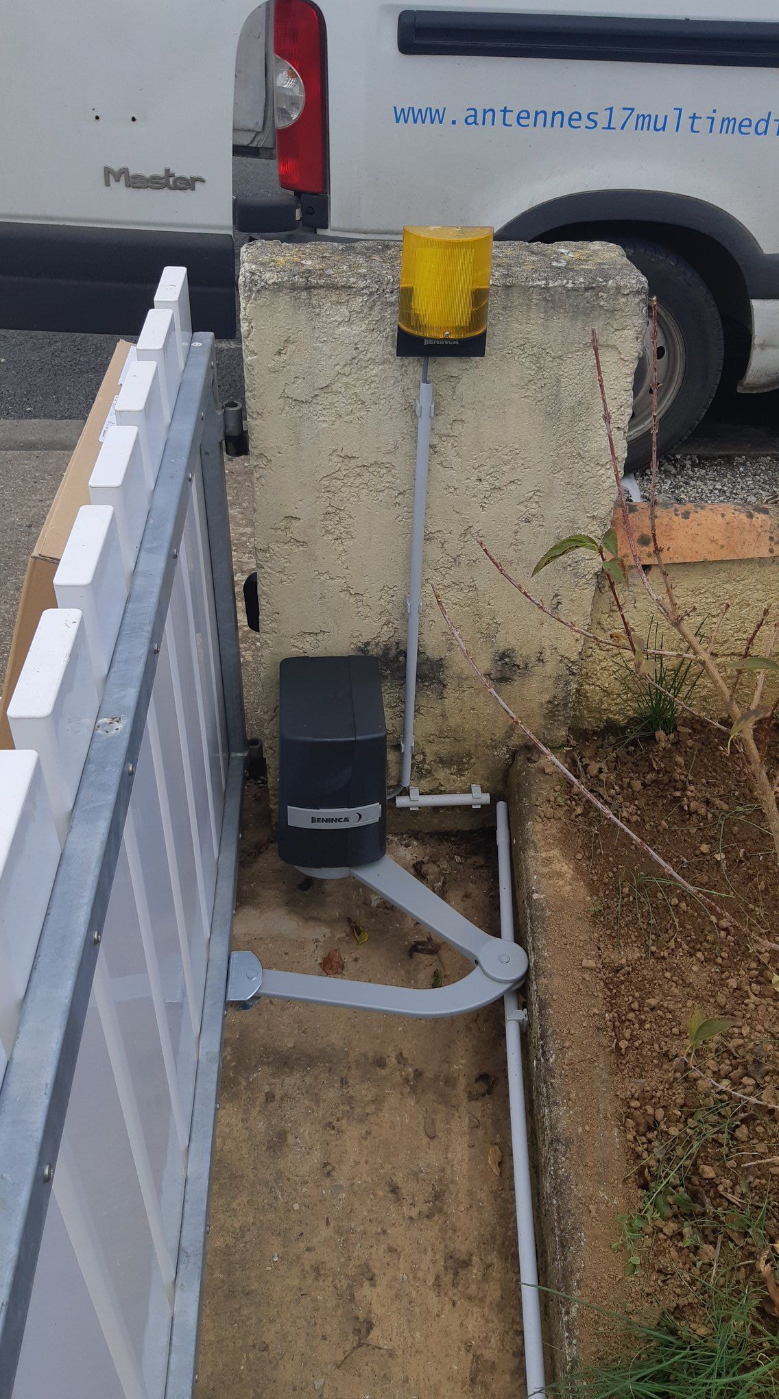 Système de portail automatique avec bras, voyant lumineux et boîtier de commande, à côté d'une clôture blanche.