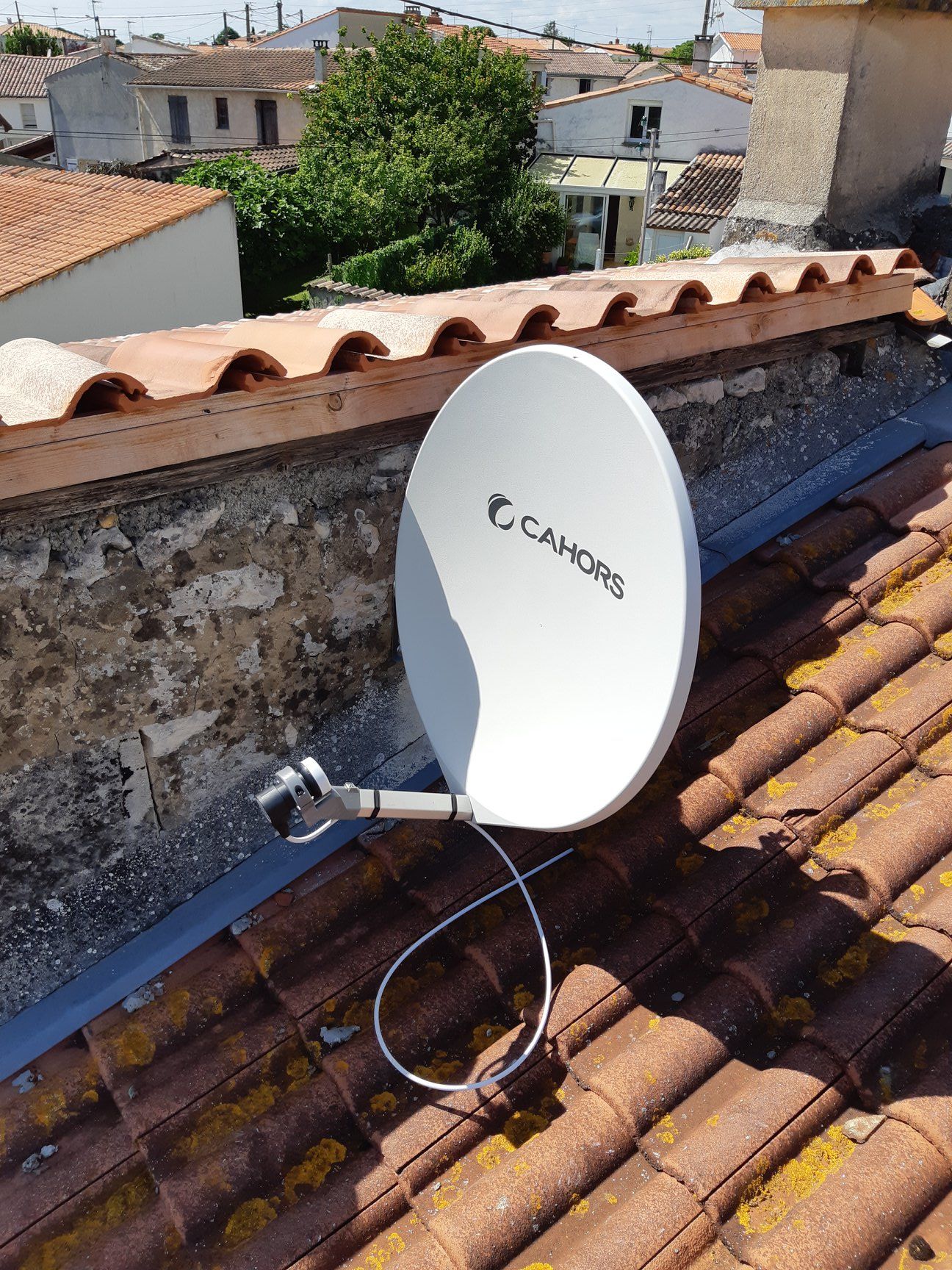Antenne parabolique blanche sur un toit en tuiles avec en arrière-plan des bâtiments et de la verdure.