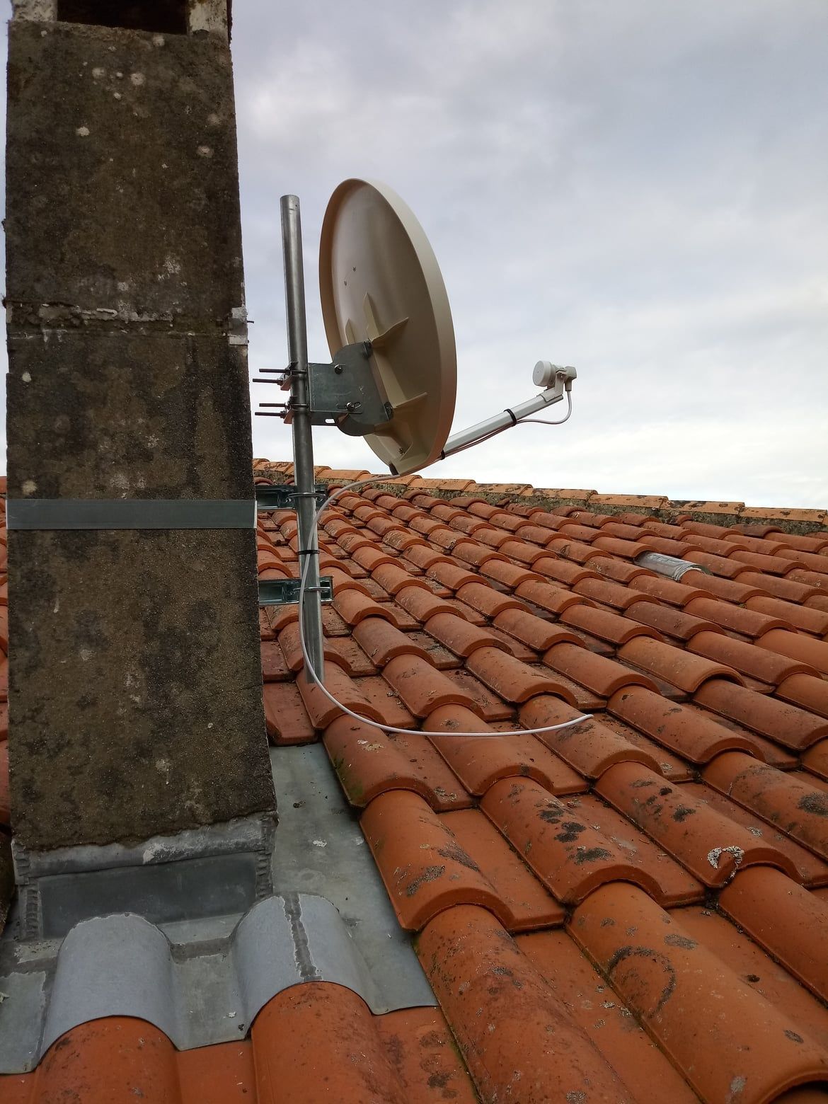 Antenne parabolique montée sur une cheminée au-dessus d'un toit en tuiles orange.