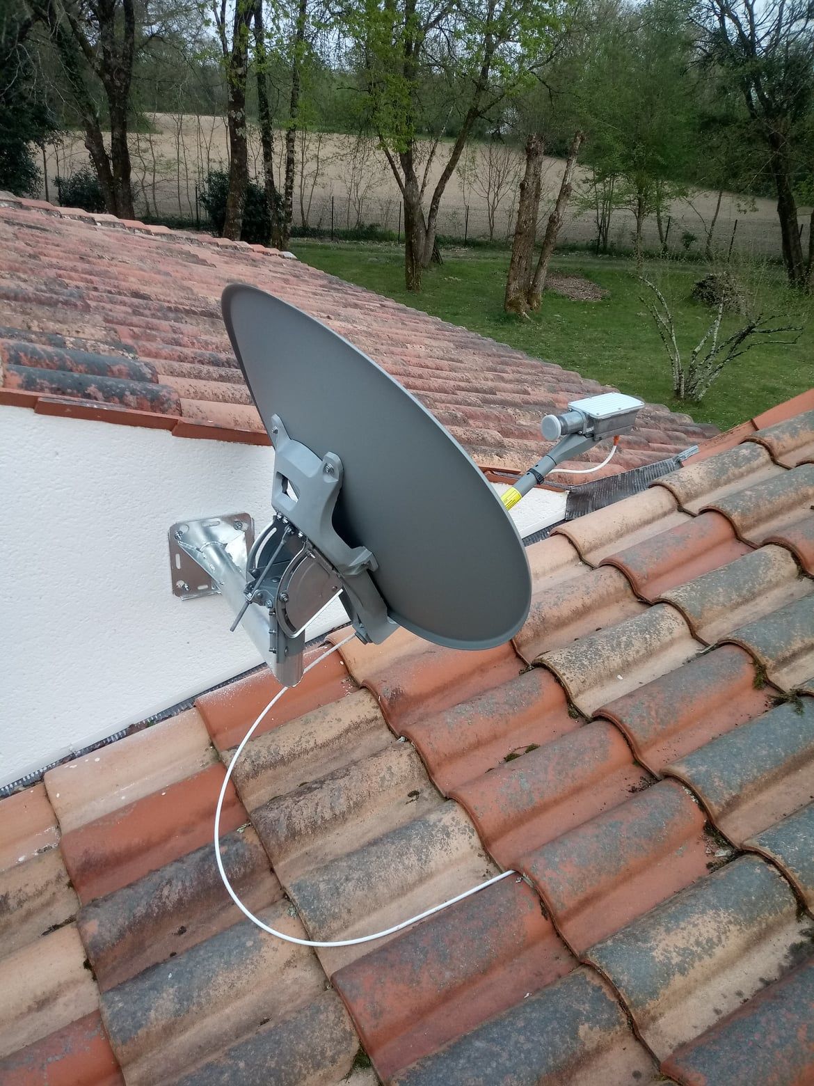 Une antenne parabolique, grise, montée sur un toit de tuiles rouges près d'un mur blanc et d'une cour avec des arbres.