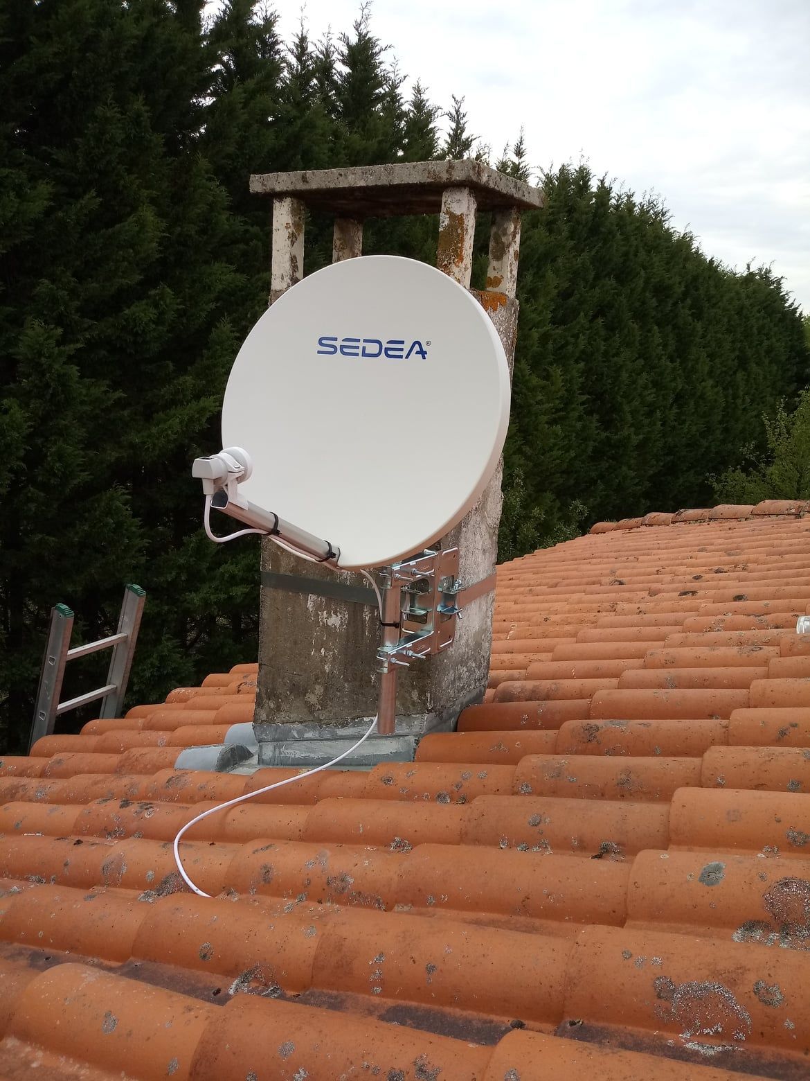 Antenne parabolique sur une cheminée, montée sur un toit de tuiles rouges. Arbres en arrière-plan.