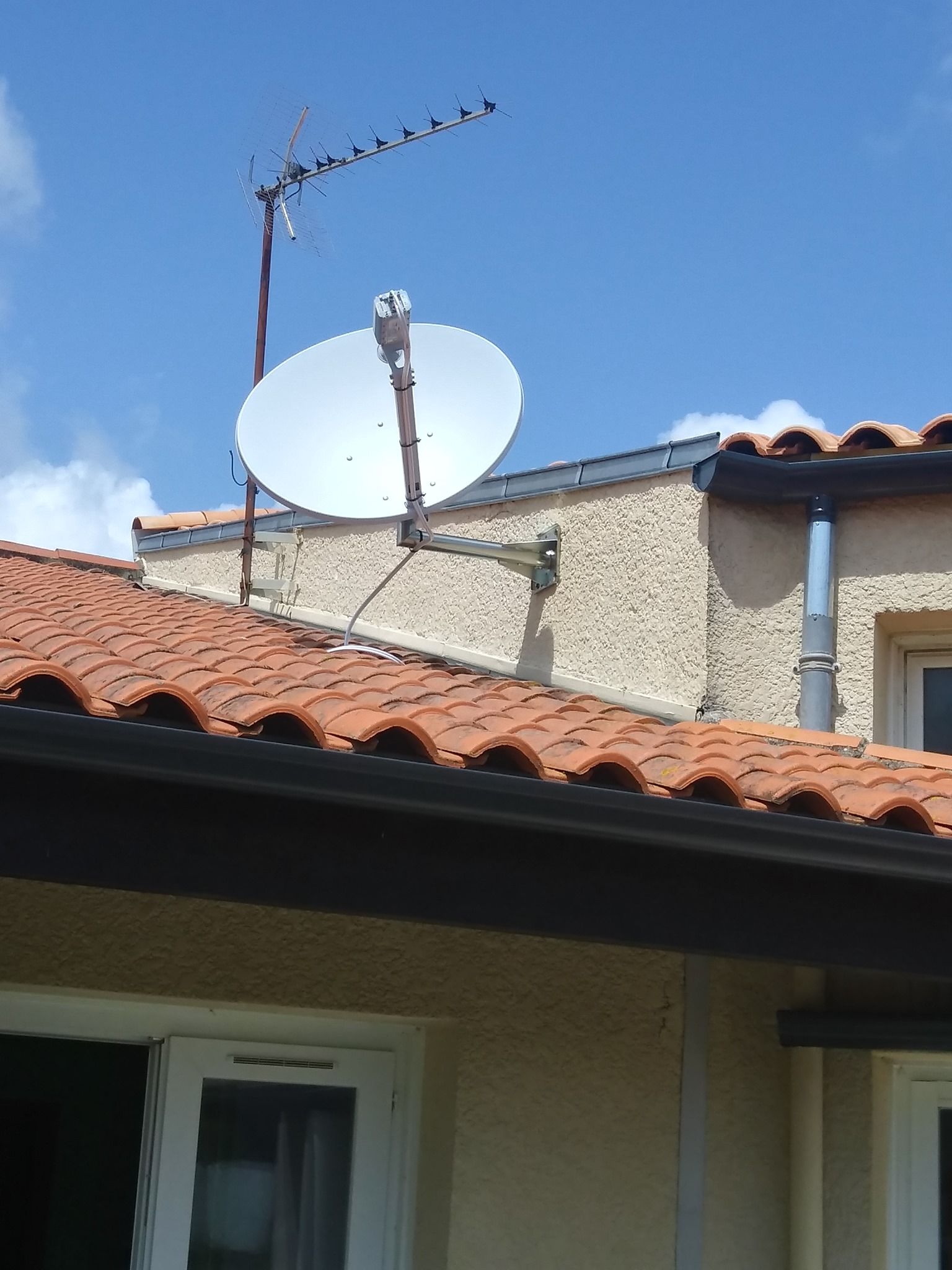 Antenne parabolique blanche et antenne sur un toit de tuiles rouges contre un ciel bleu.