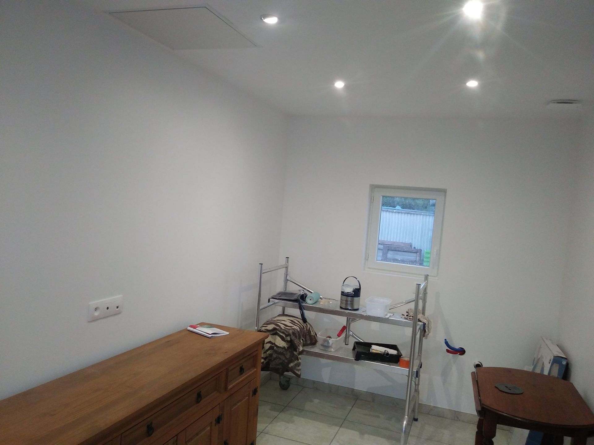 Pièce aux murs blancs avec une commode en bois, des étagères en métal, une petite fenêtre et une table d'appoint.